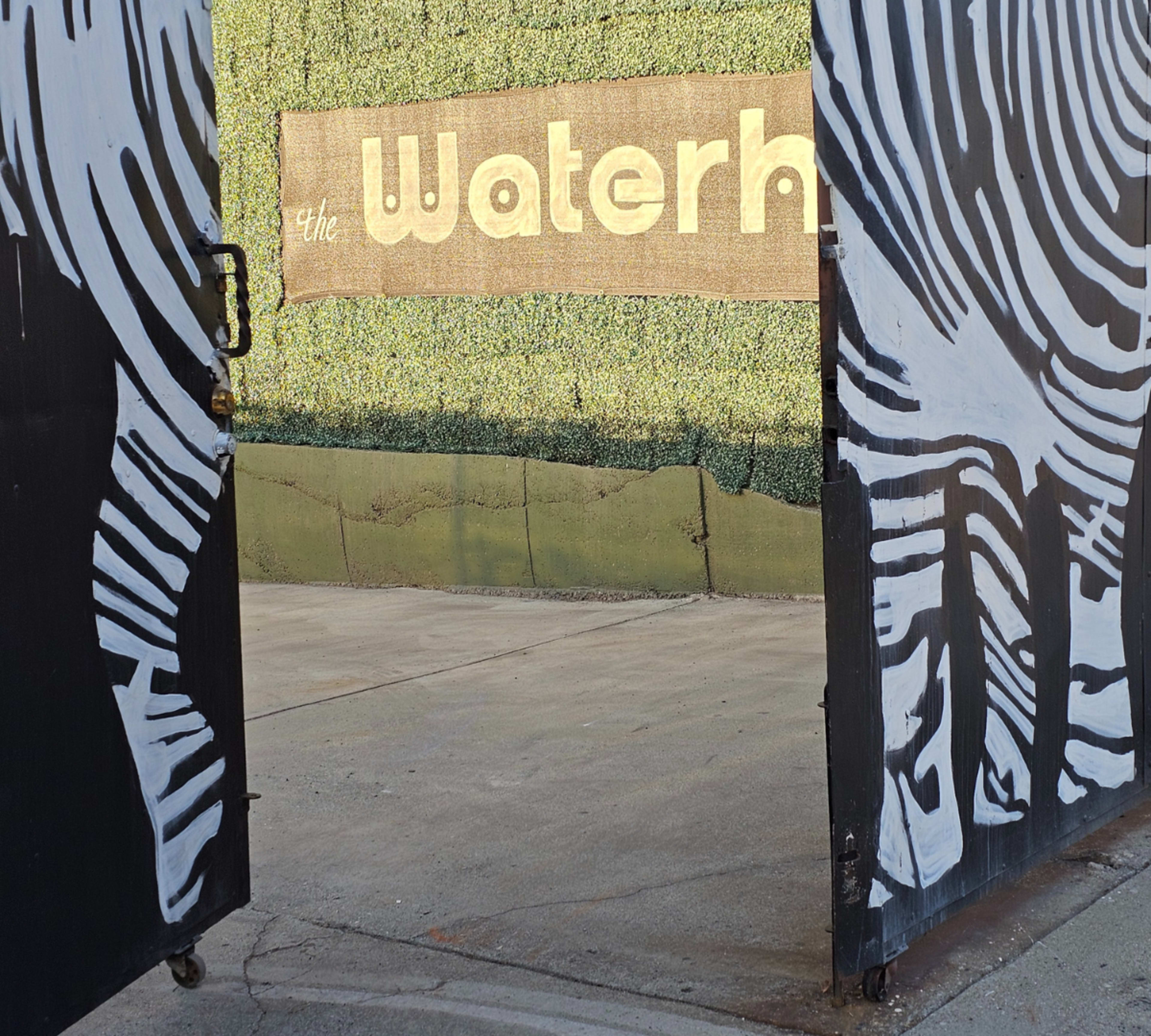An open gate with zebra-striped doors, revealing a sign that reads "the Waterh." against a green hedge backdrop.