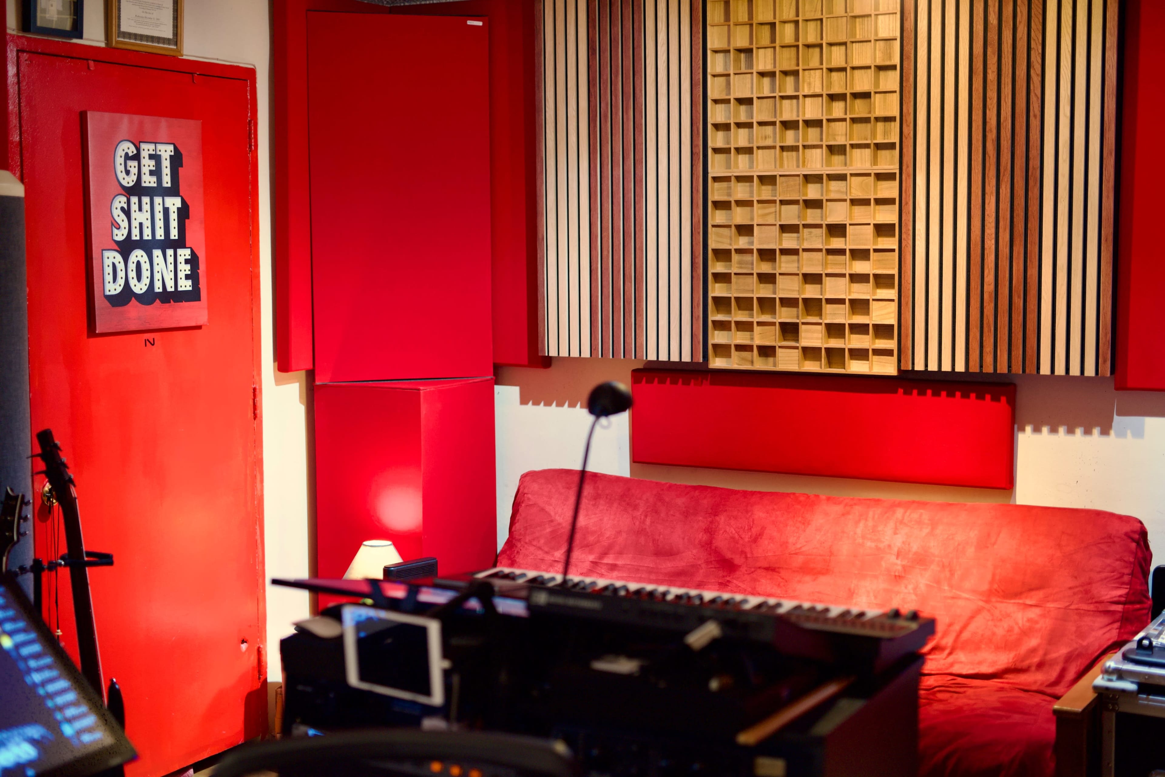 A music studio with red soundproofing panels, a wooden sound diffuser, a red couch, and various musical equipment in the foreground.