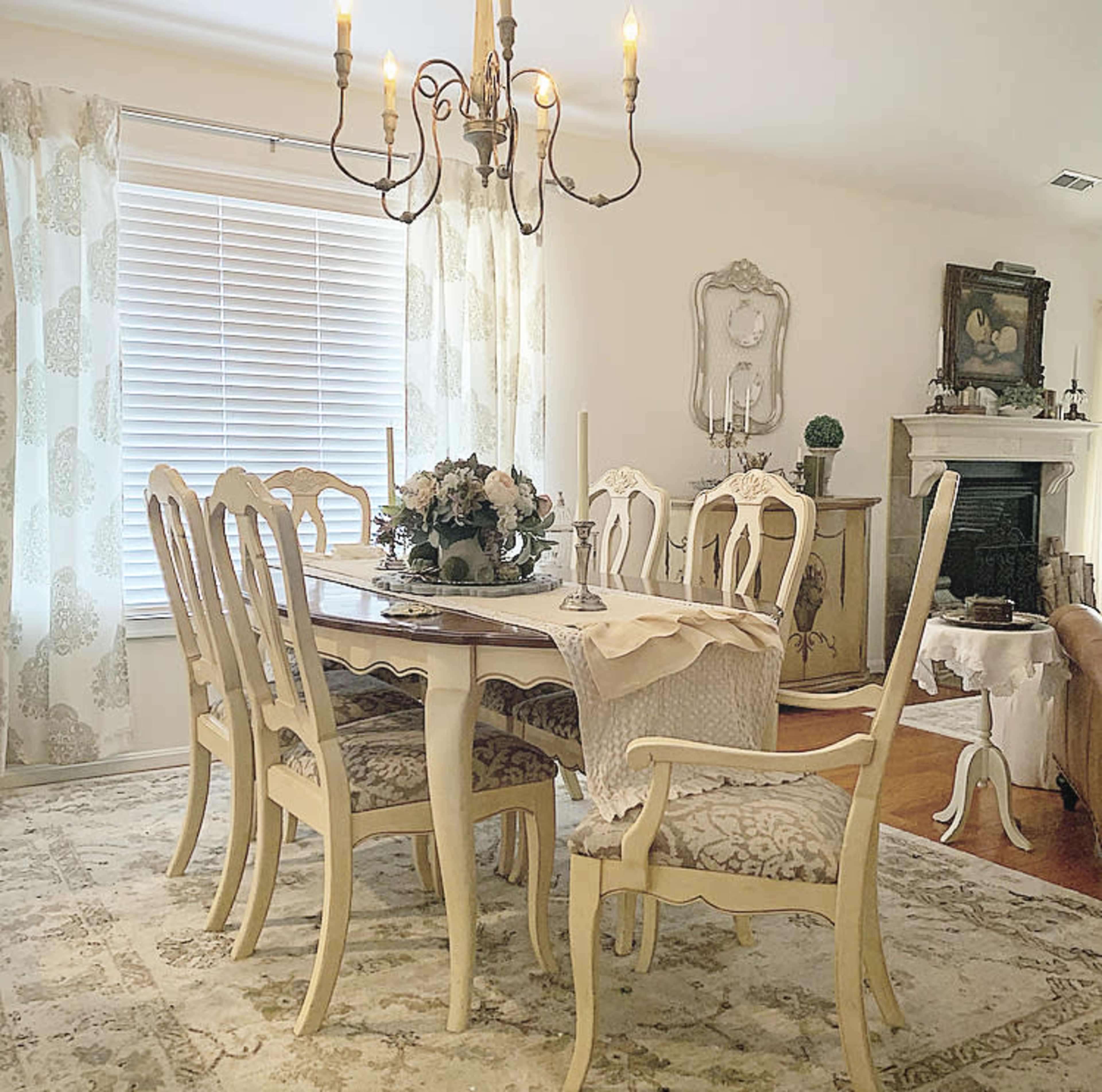 A dining room features a wooden table surrounded by six chairs, with a chandelier overhead and floral decorations on the table.