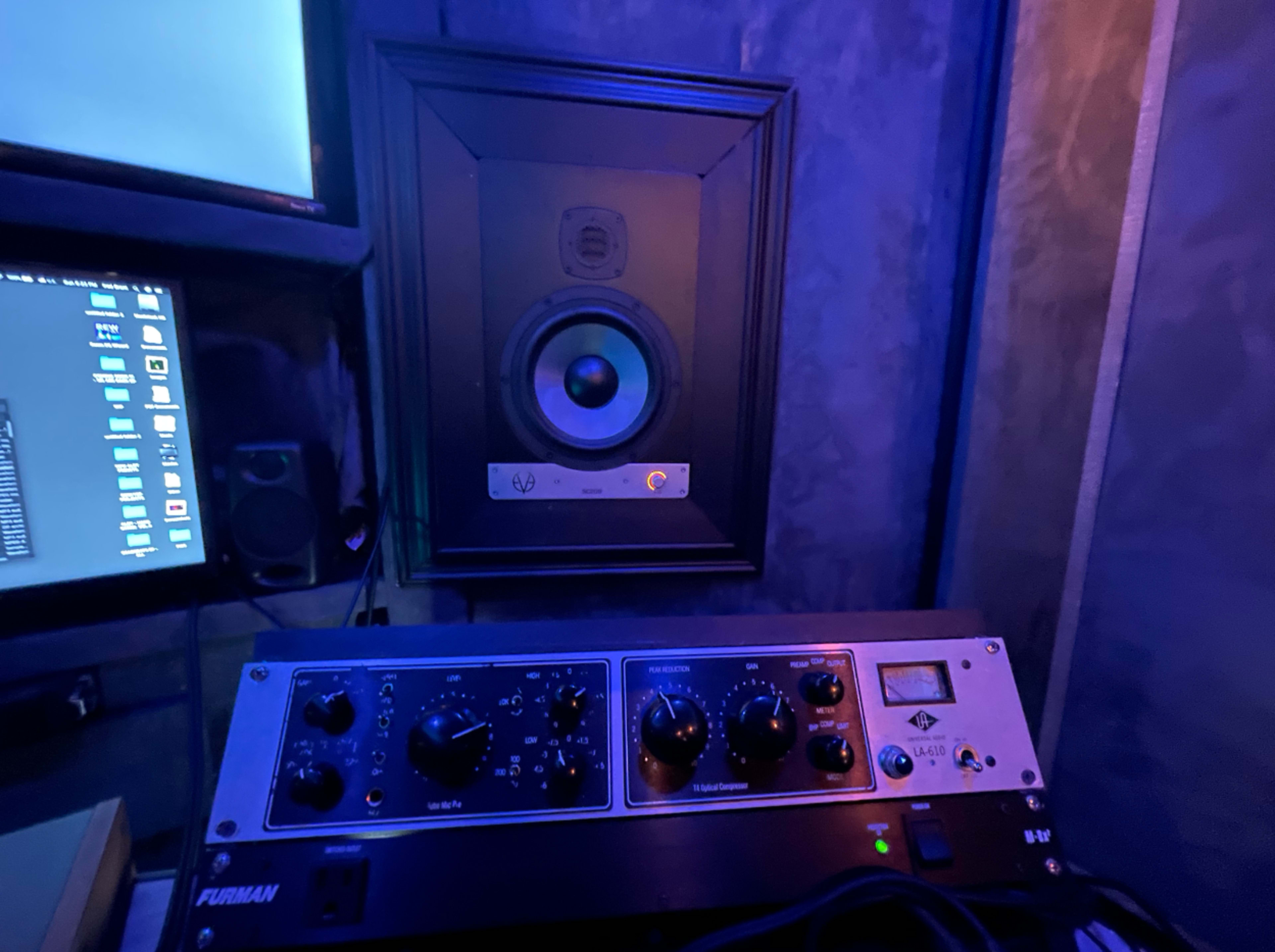 The image shows a studio setup with a speaker mounted on the wall above an audio processing unit featuring various knobs and controls.