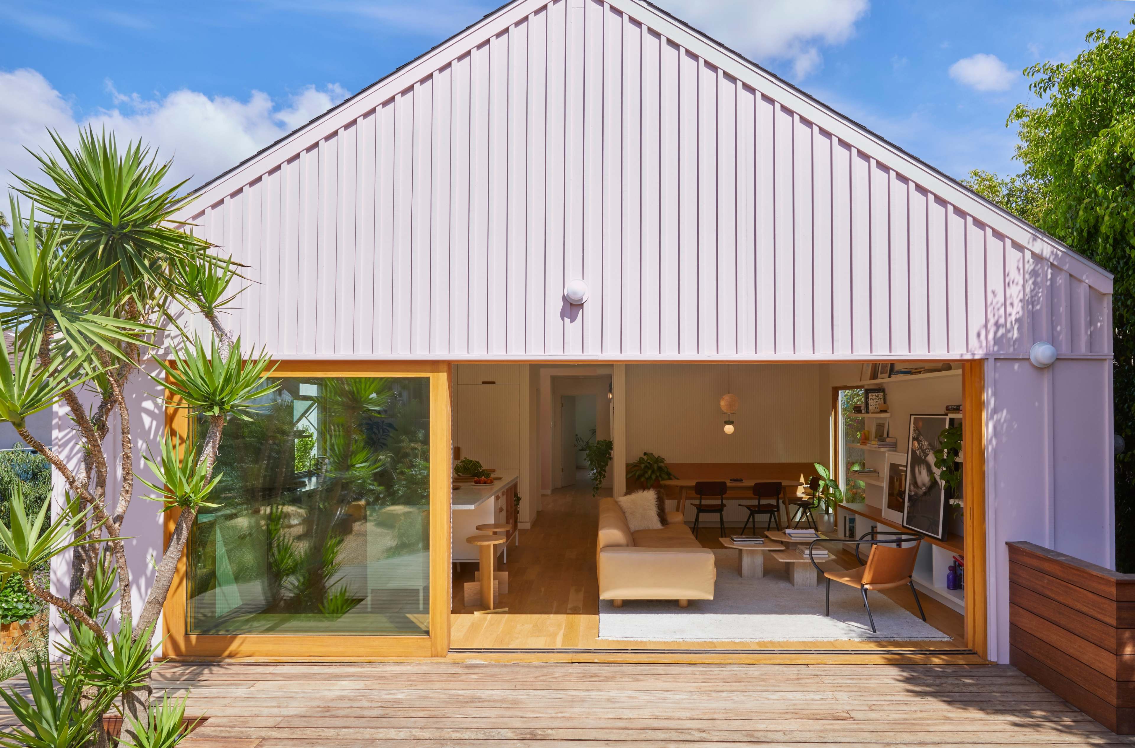 A modern home with a pink metal roof, large glass doors opening to a wooden deck, and surrounding tropical plants.