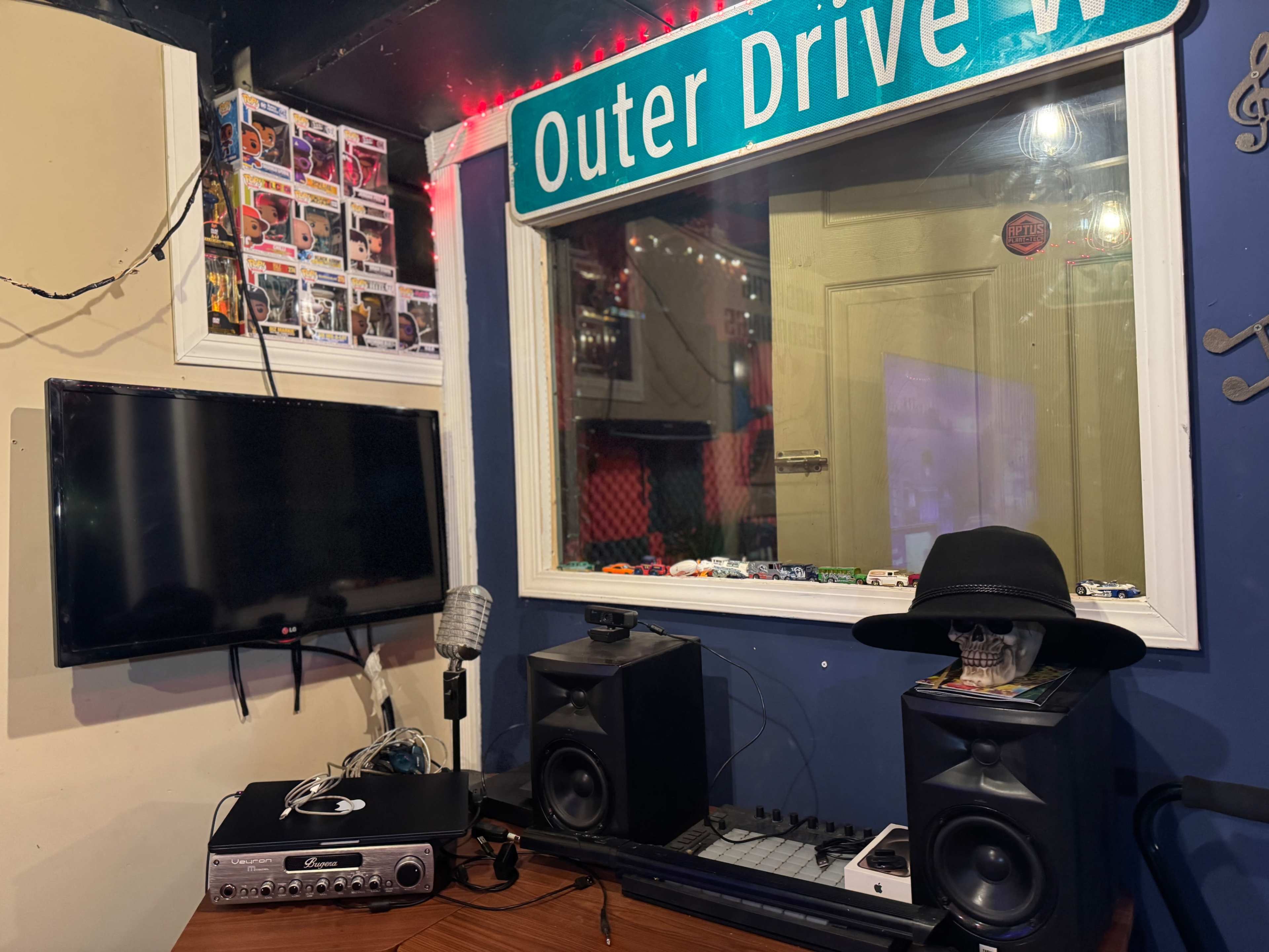 The image shows a room with a wooden table featuring audio equipment, a television, and a window covered with a screen, while a green street sign labeled "Outer Drive W" hangs above.