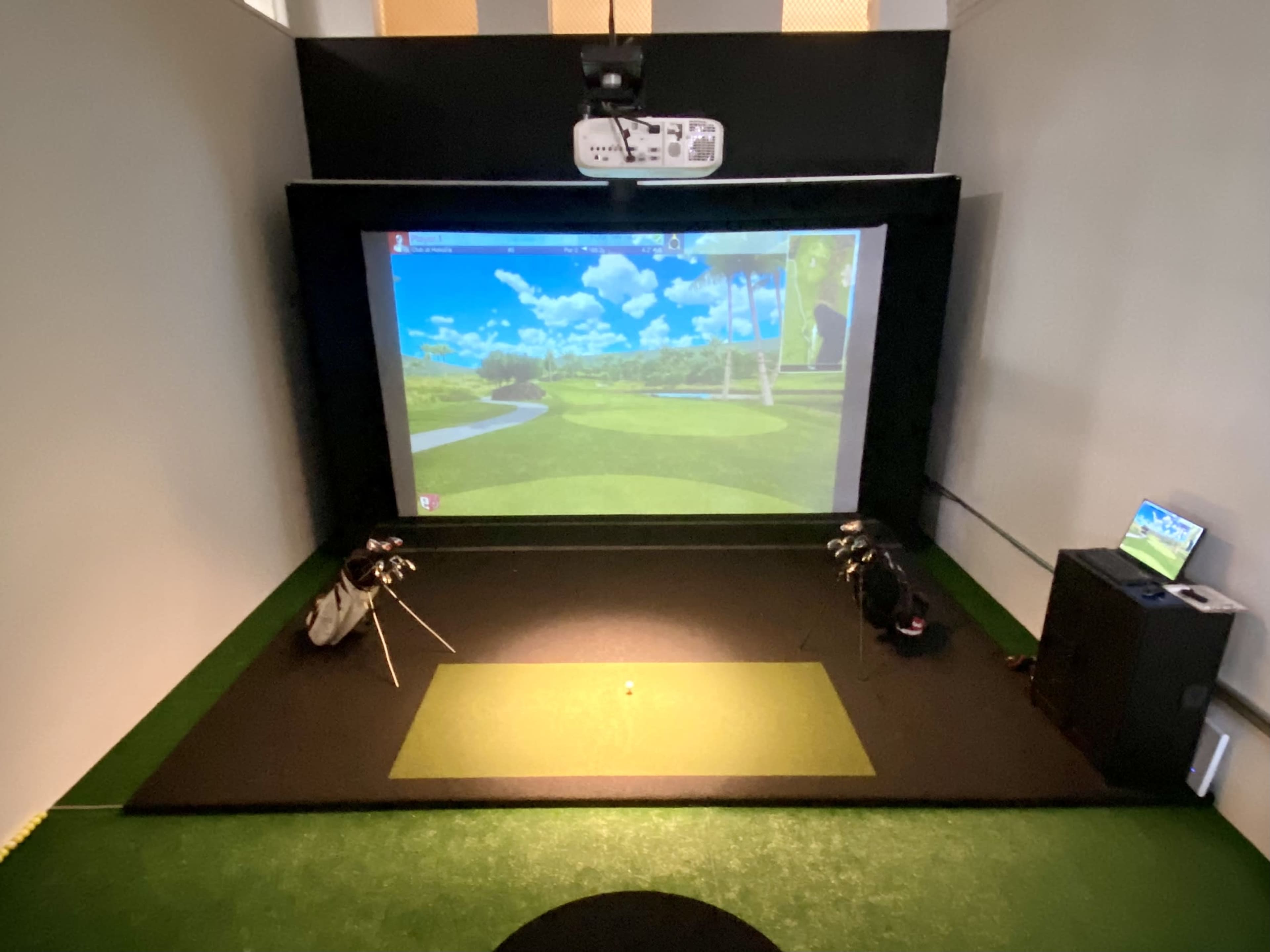 An indoor golf simulator setup with a large screen displaying a golf course, a putting green, and golf bags positioned on the floor.
