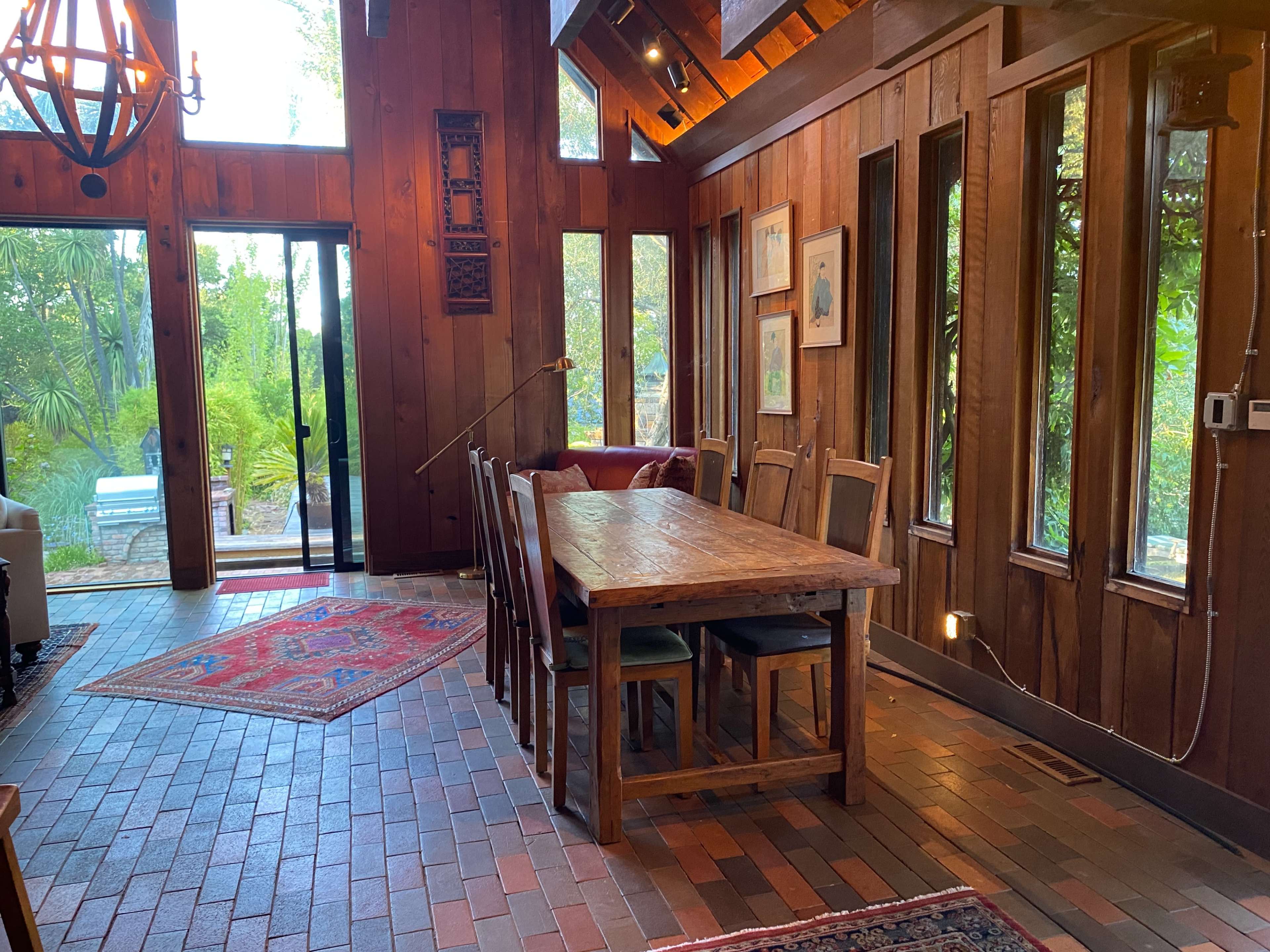 A wooden dining area with a large table and chairs, illuminated by natural light from surrounding windows, next to a patio door leading to an outdoor space.