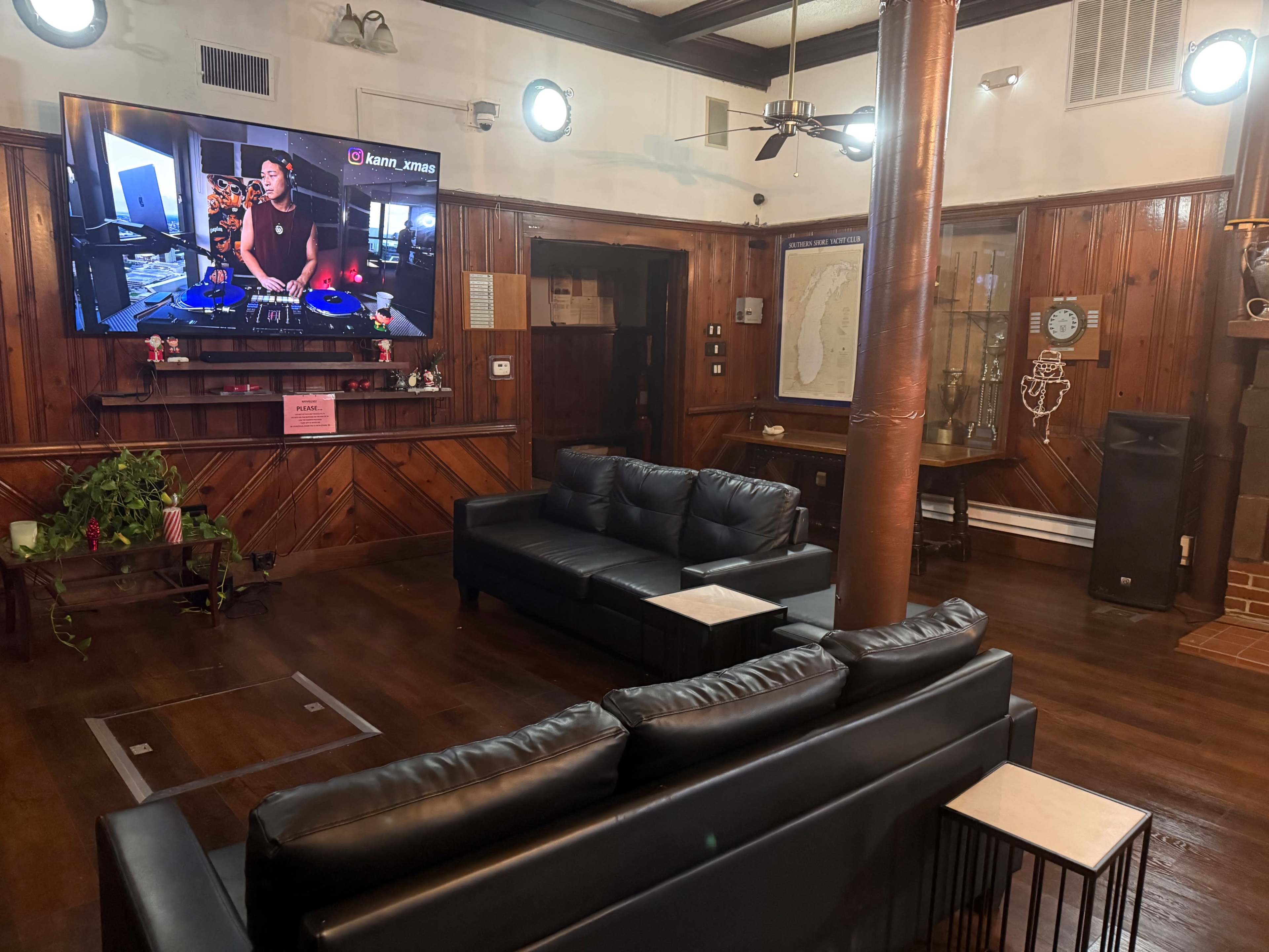 The image shows a lounge area with black sofas facing a large television displaying a person DJing, surrounded by wooden paneling and a decorative fireplace.