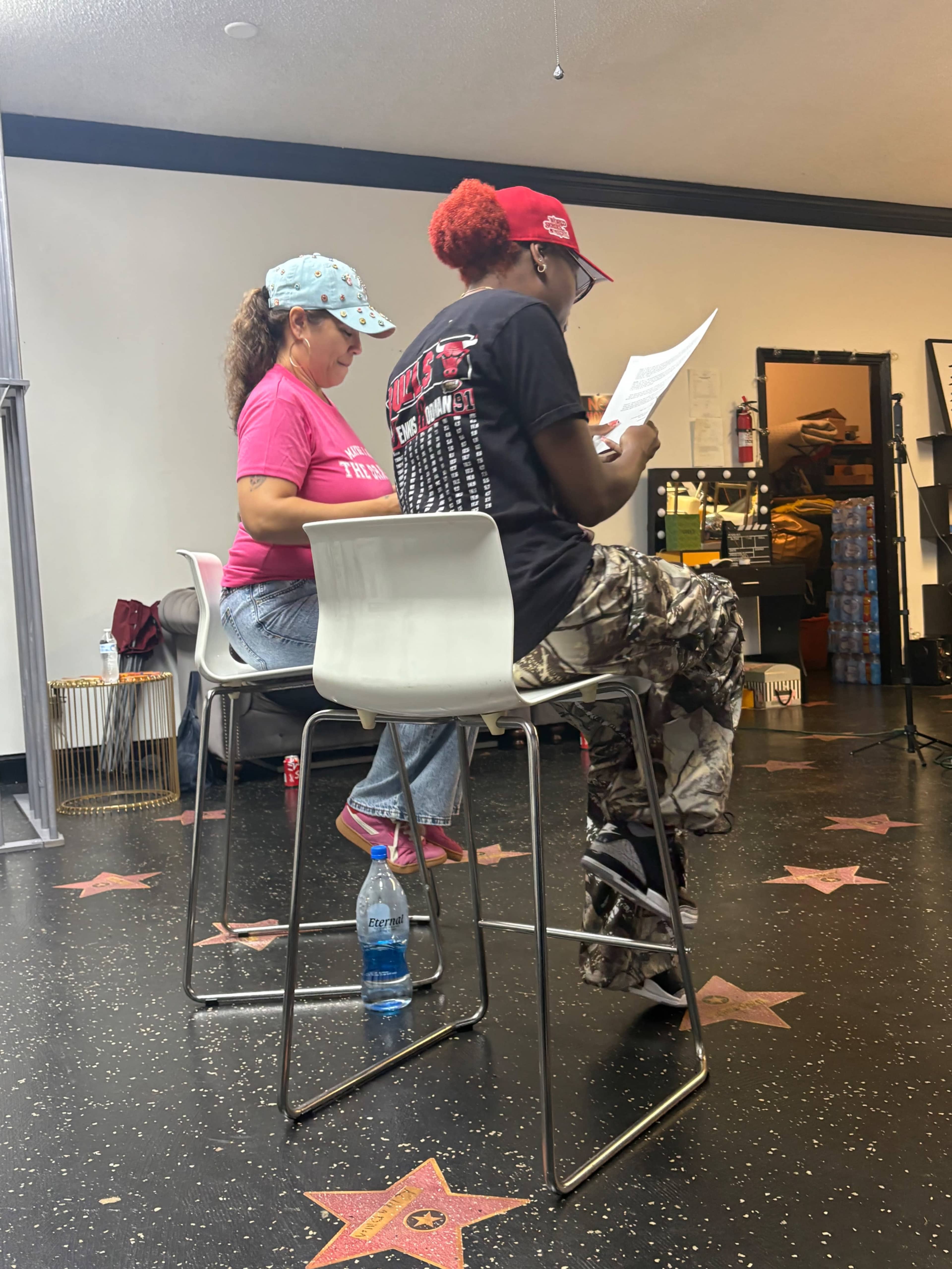 Two individuals sit on high chairs in a rehearsal space, reading from papers in front of them.