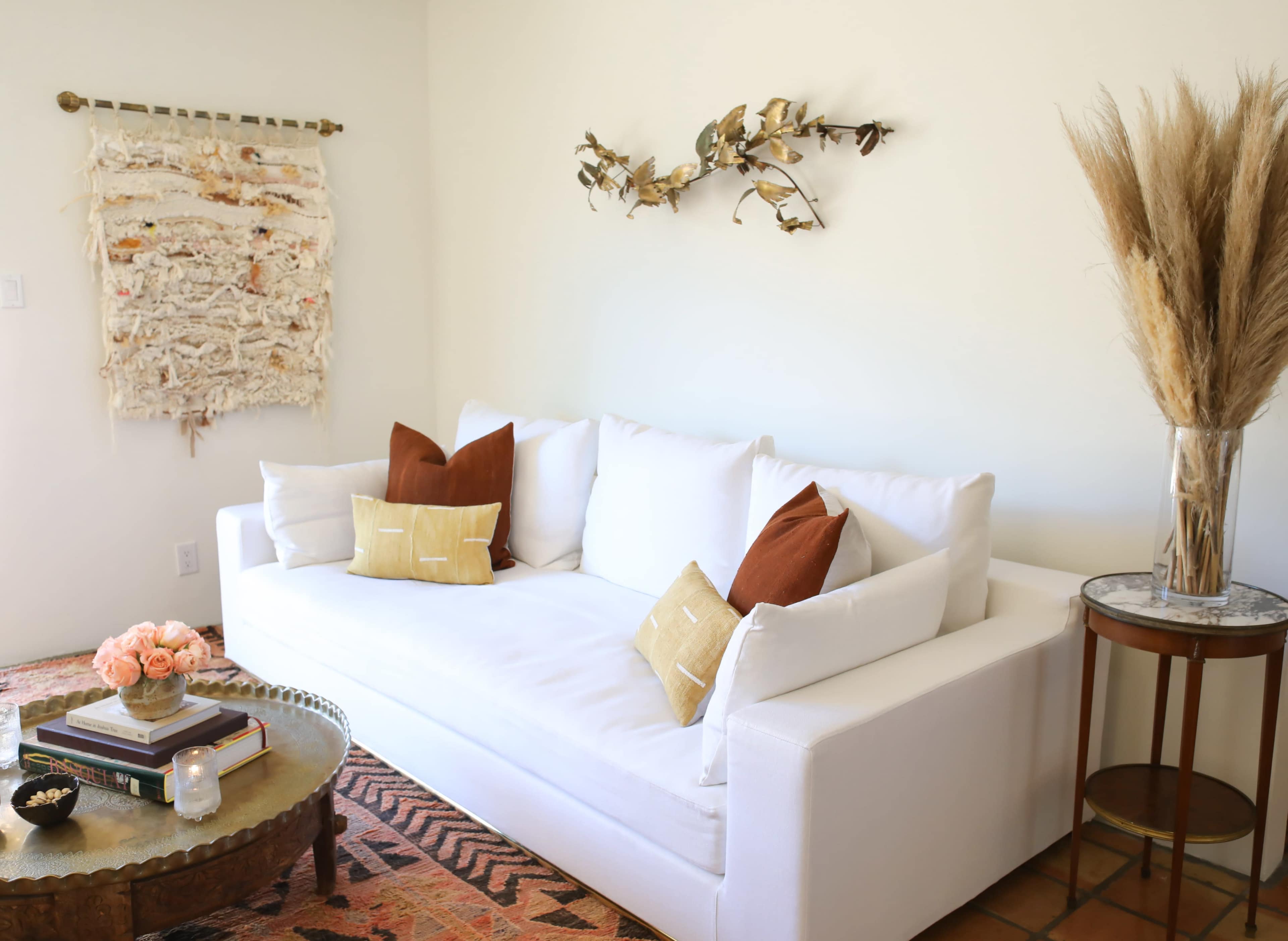 A bright, minimalist living room featuring a white sofa with decorative cushions, a round wooden coffee table, and a woven wall hanging.