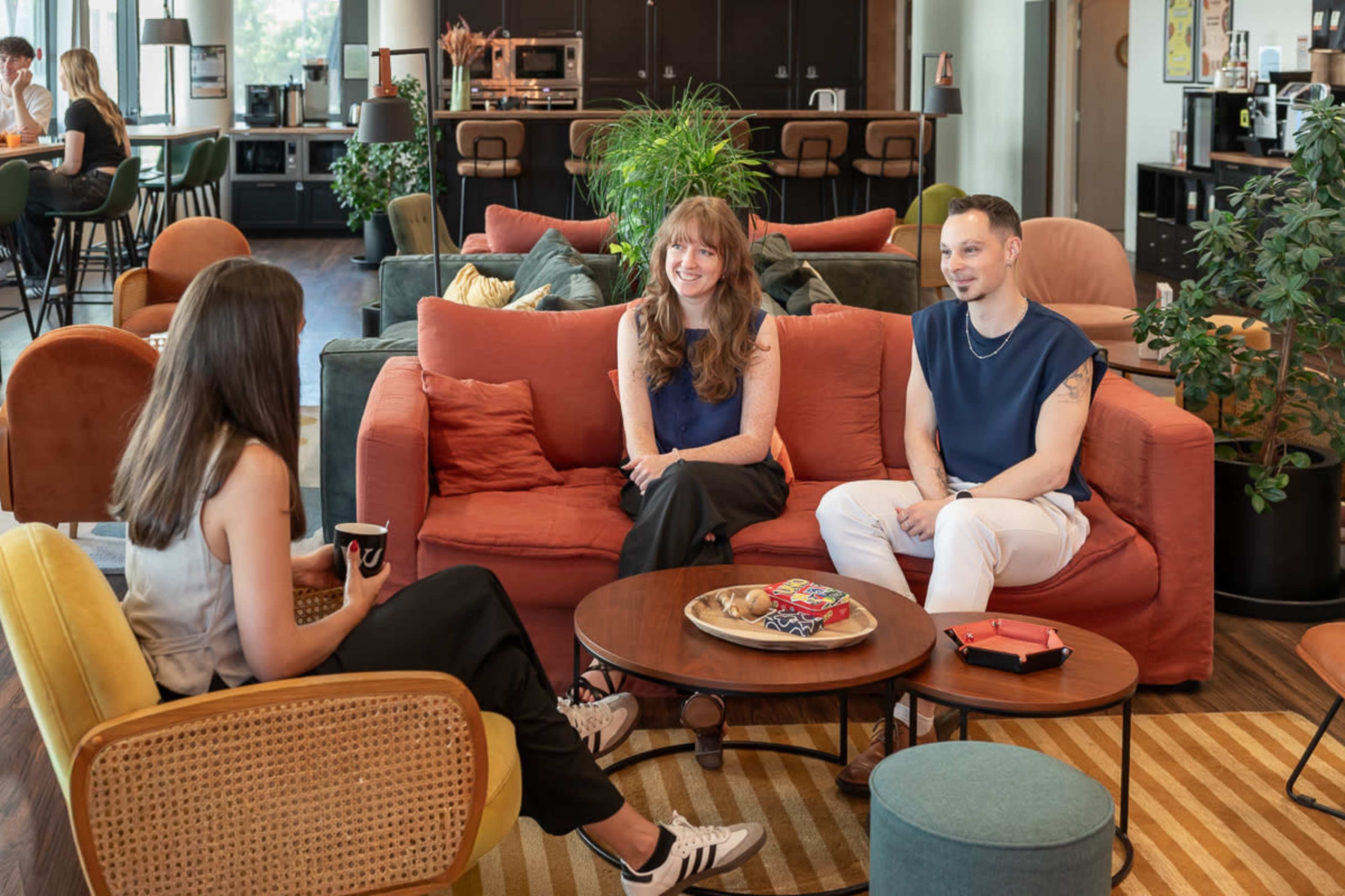 Three individuals are seated in a cozy lounge area with a mix of sofas and chairs, engaged in conversation over coffee amid greenery and a casual atmosphere.