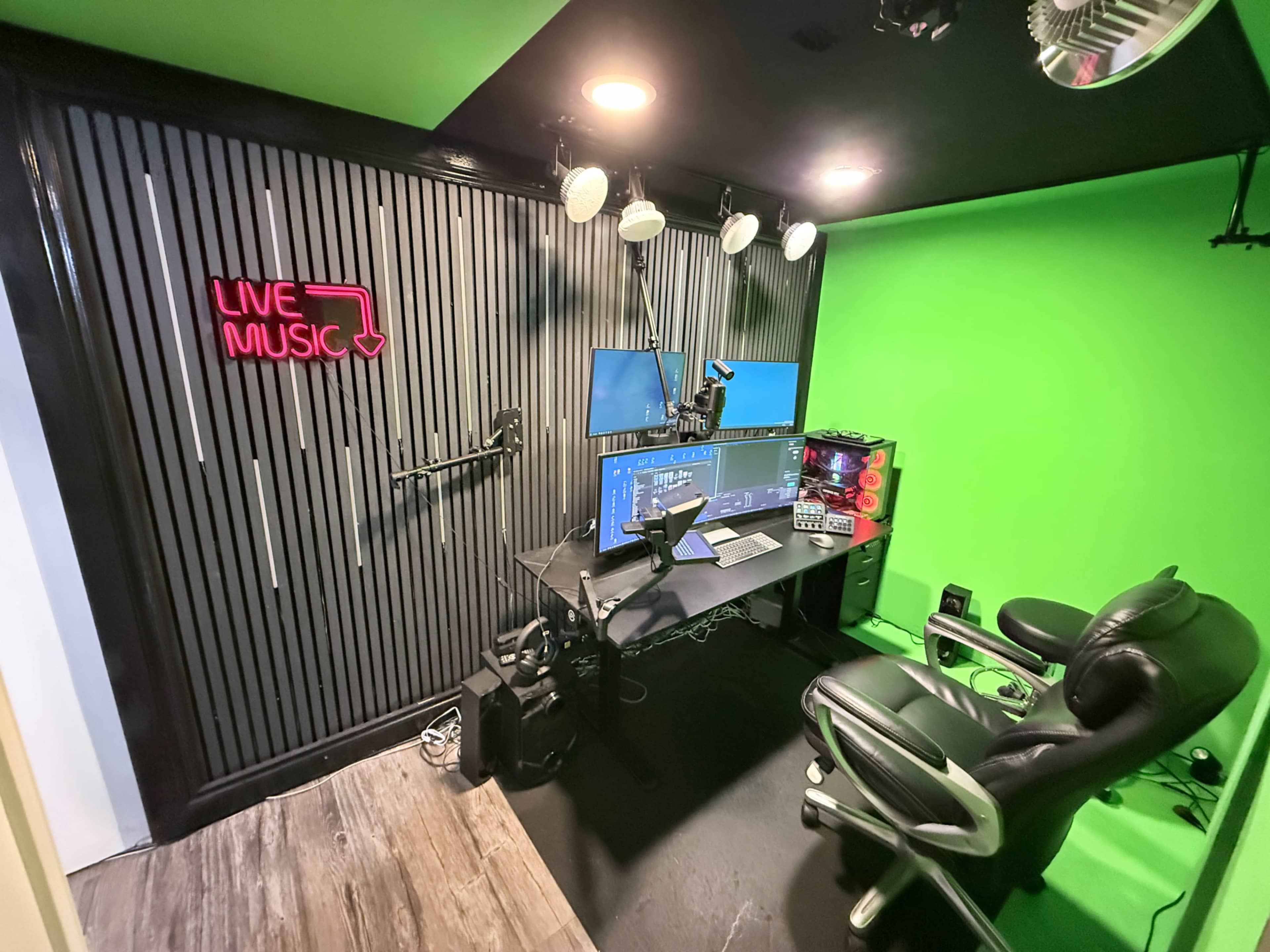 The image shows a compact media studio with a green screen backdrop, multiple computer monitors, and a black desk featuring a microphone and various audio equipment.