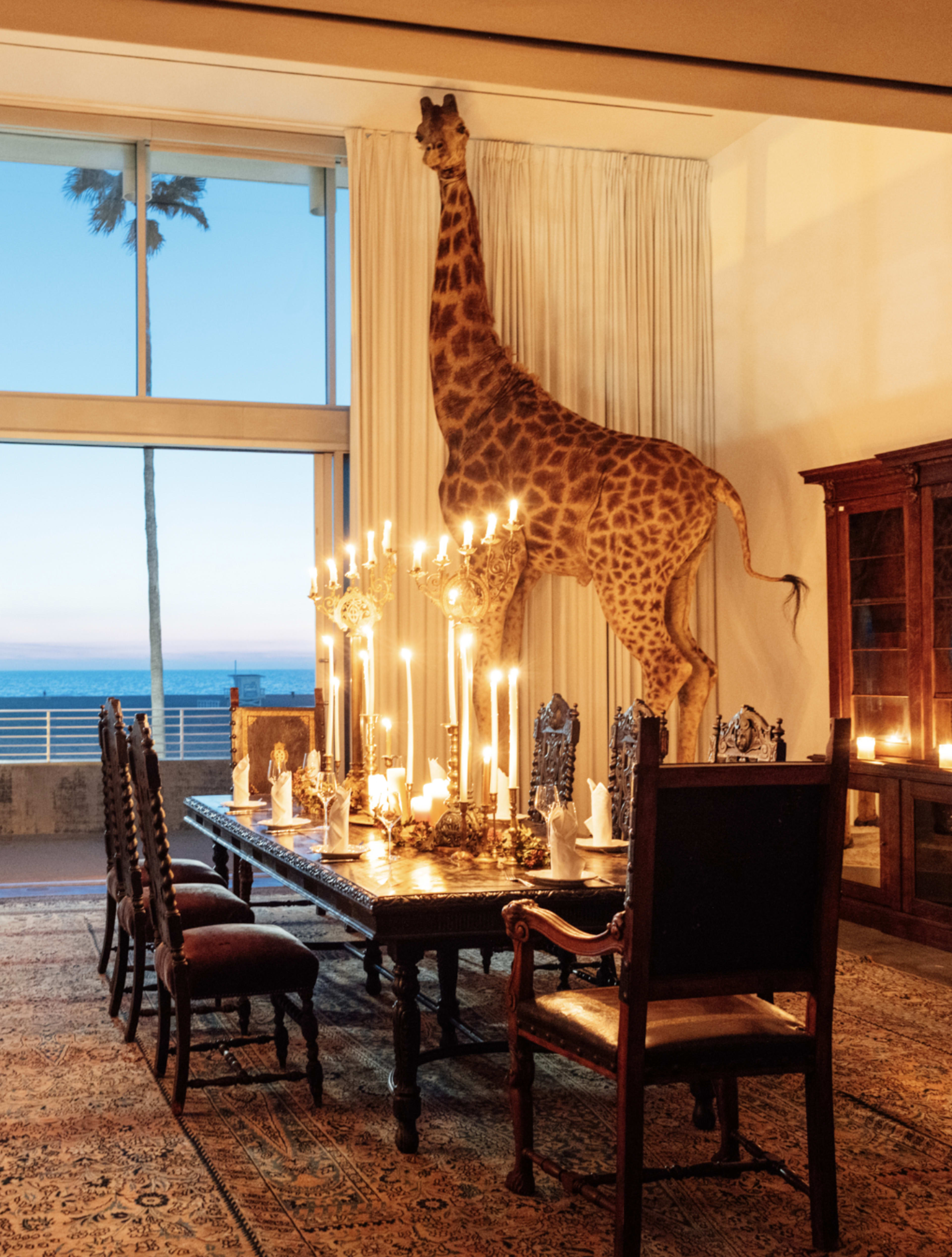 A giraffe sculpture is mounted against the wall above a dining table set with candles in a room with large windows overlooking the ocean.