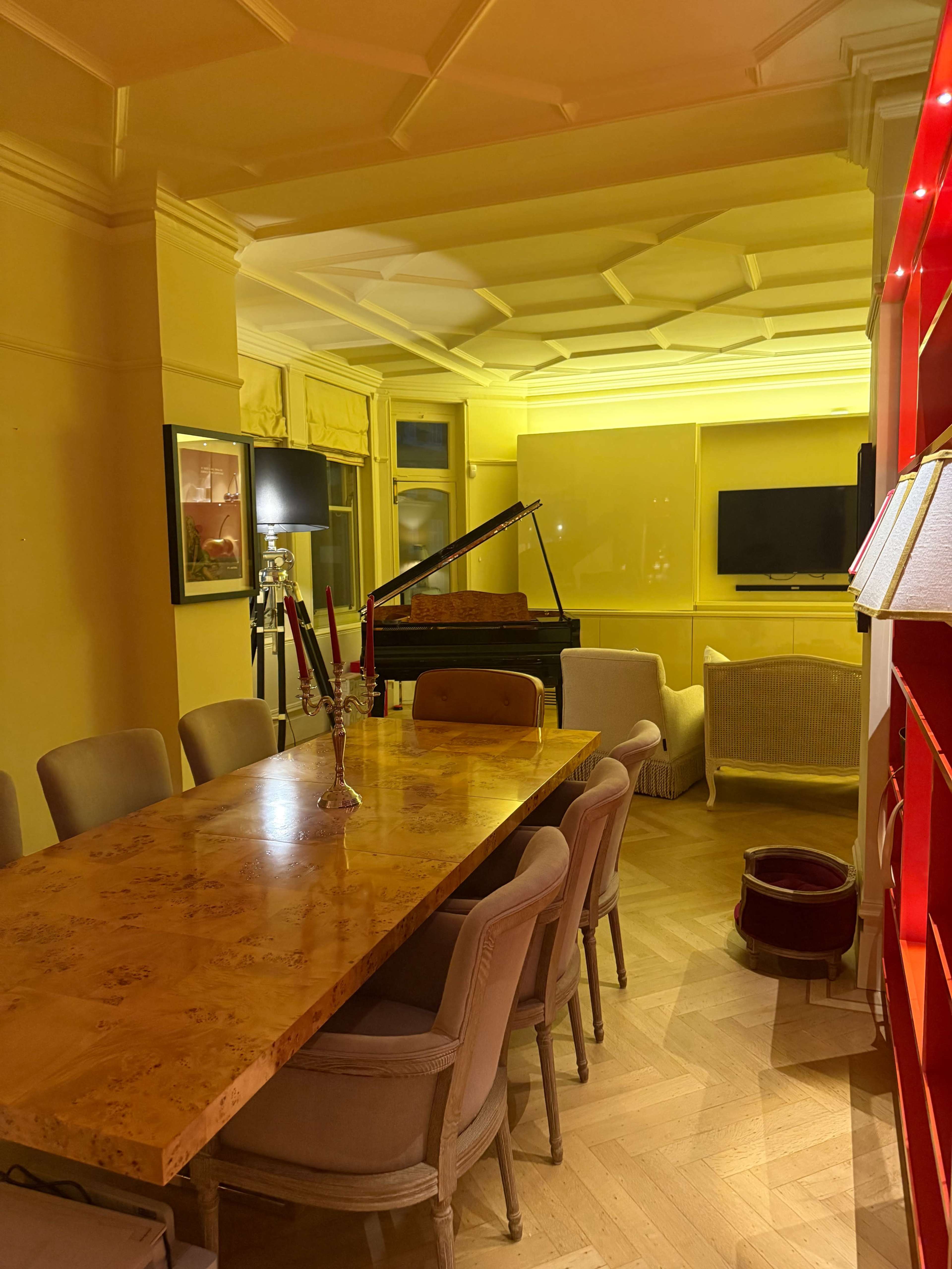 The image shows a warmly lit dining room featuring a long wooden table surrounded by upholstered chairs, a grand piano in the background, and a television mounted on the wall.