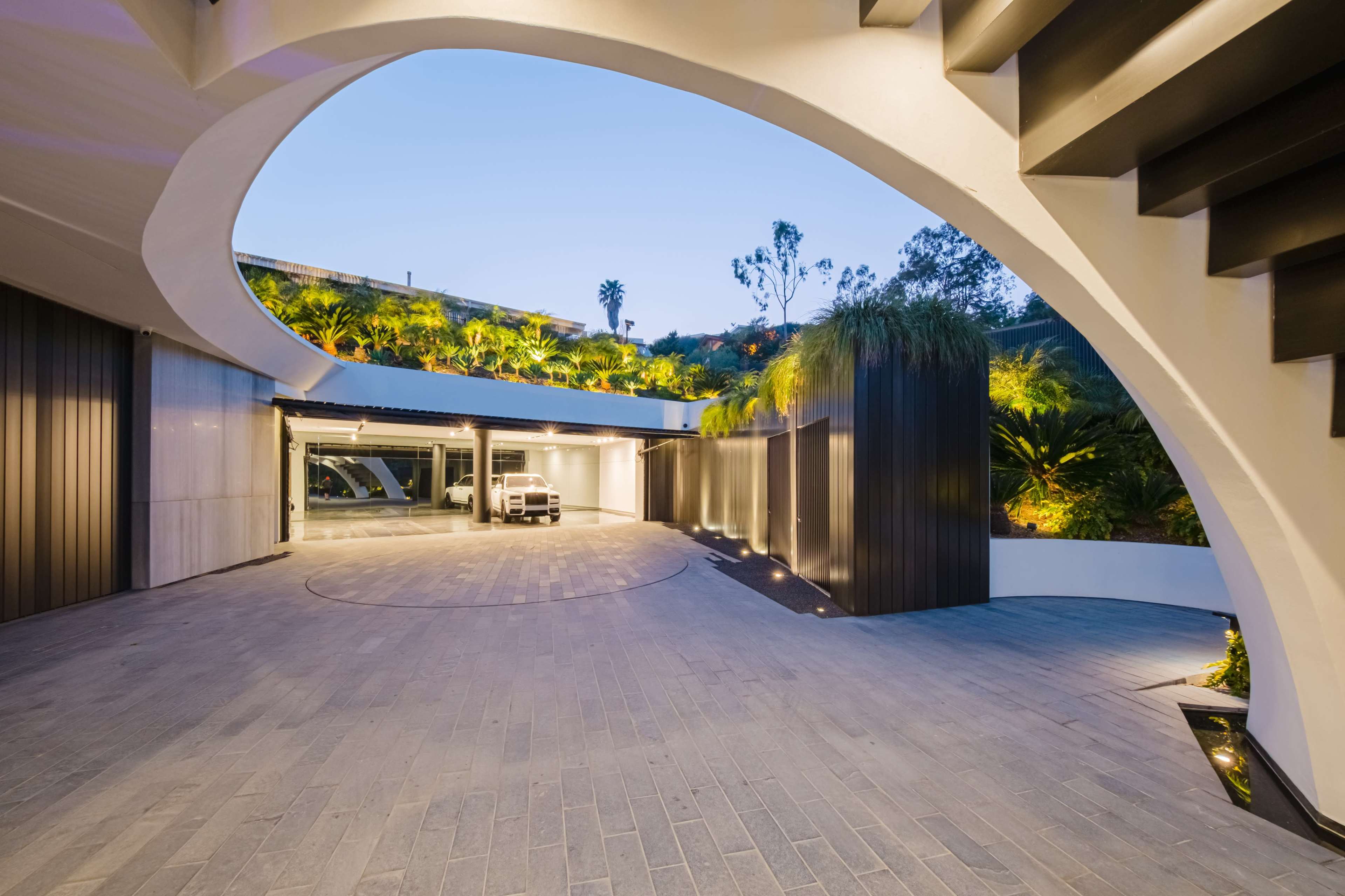 A modern architectural entrance with a curved driveway leading to an expansive garage area, surrounded by greenery and illuminated by soft lighting.