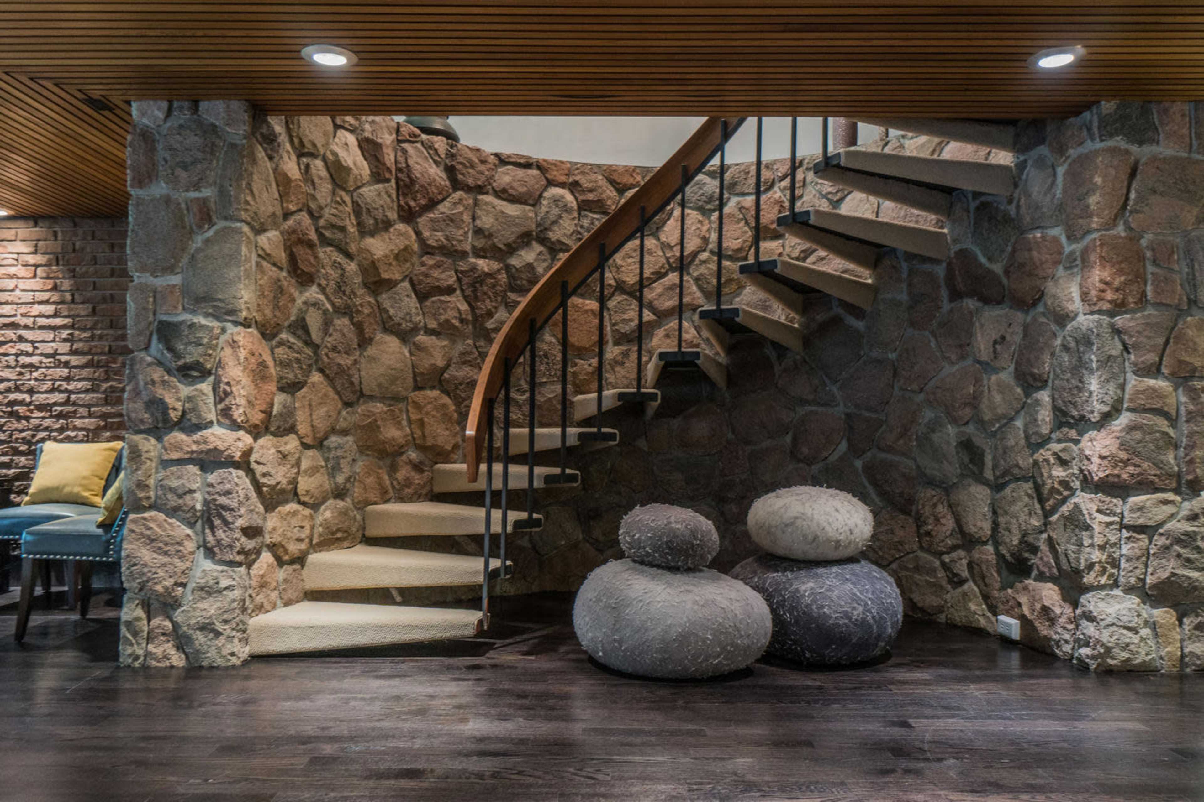 A curved staircase with wooden steps is situated next to a stone wall, alongside two round, textured stone sculptures on a dark wooden floor.