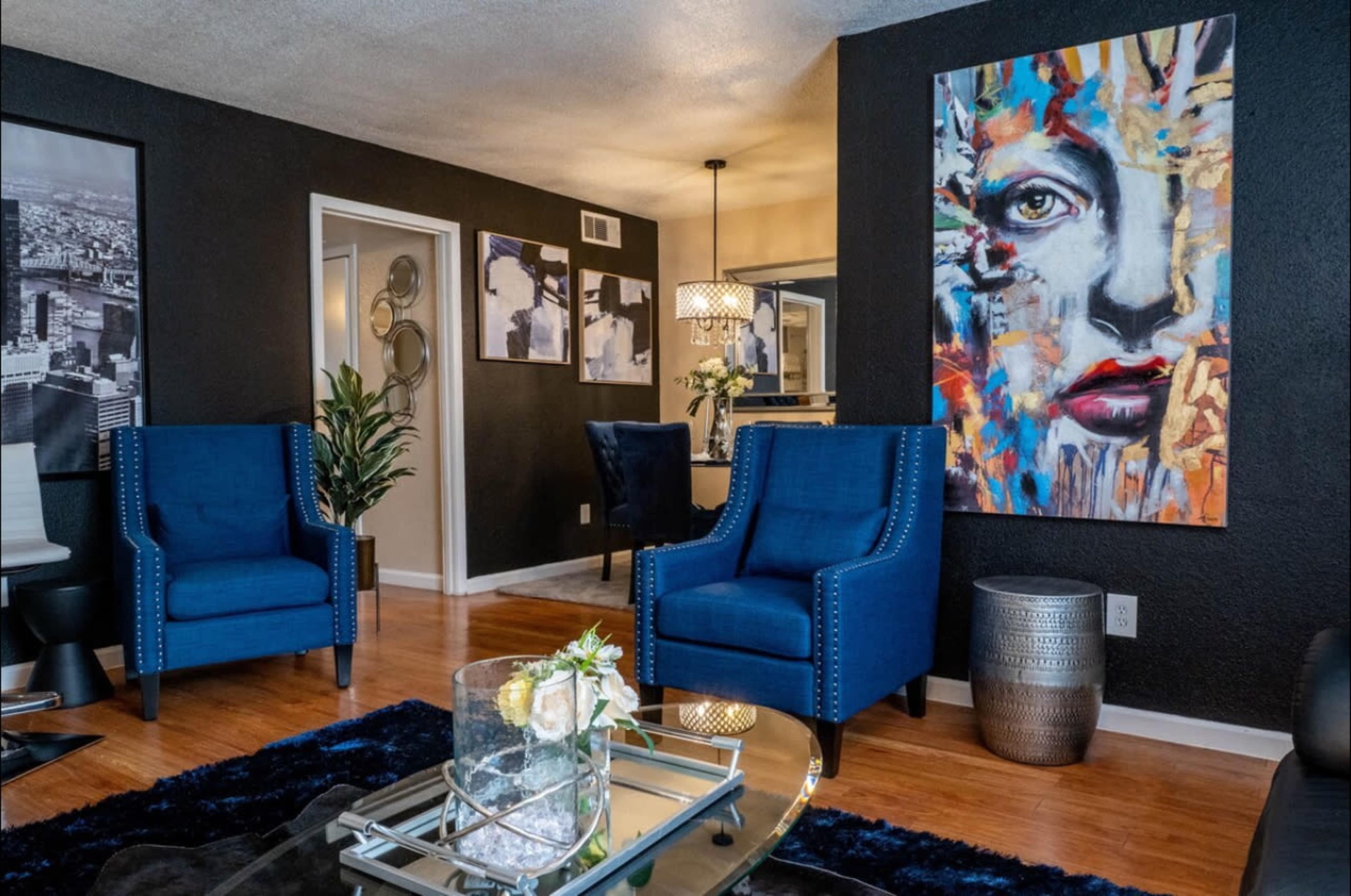 The image features a modern living room with two blue chairs, a glass coffee table, and a colorful abstract painting of a face on a black wall.