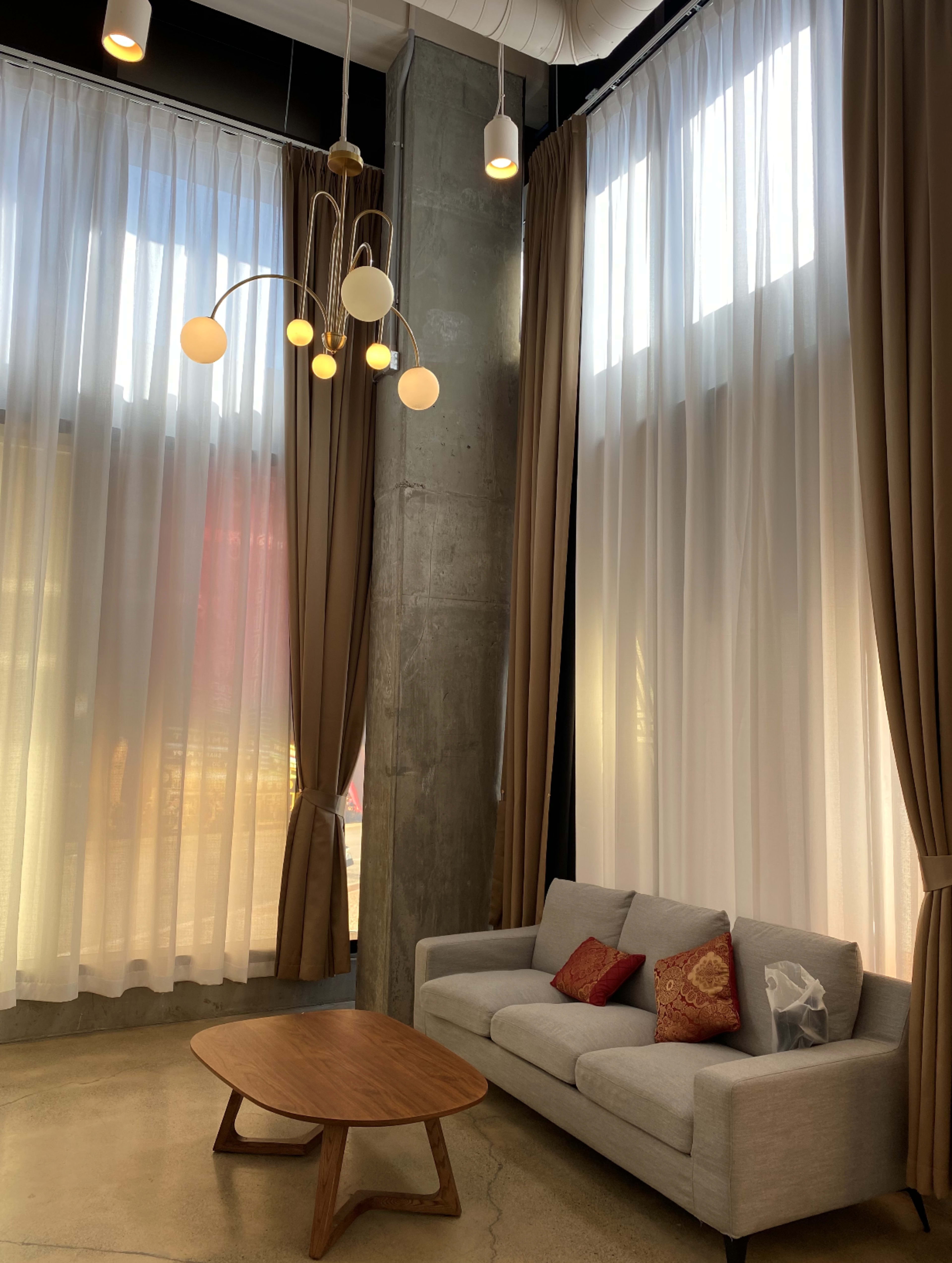 A modern living space with a gray sofa, a wooden coffee table, and large windows adorned with sheer curtains.