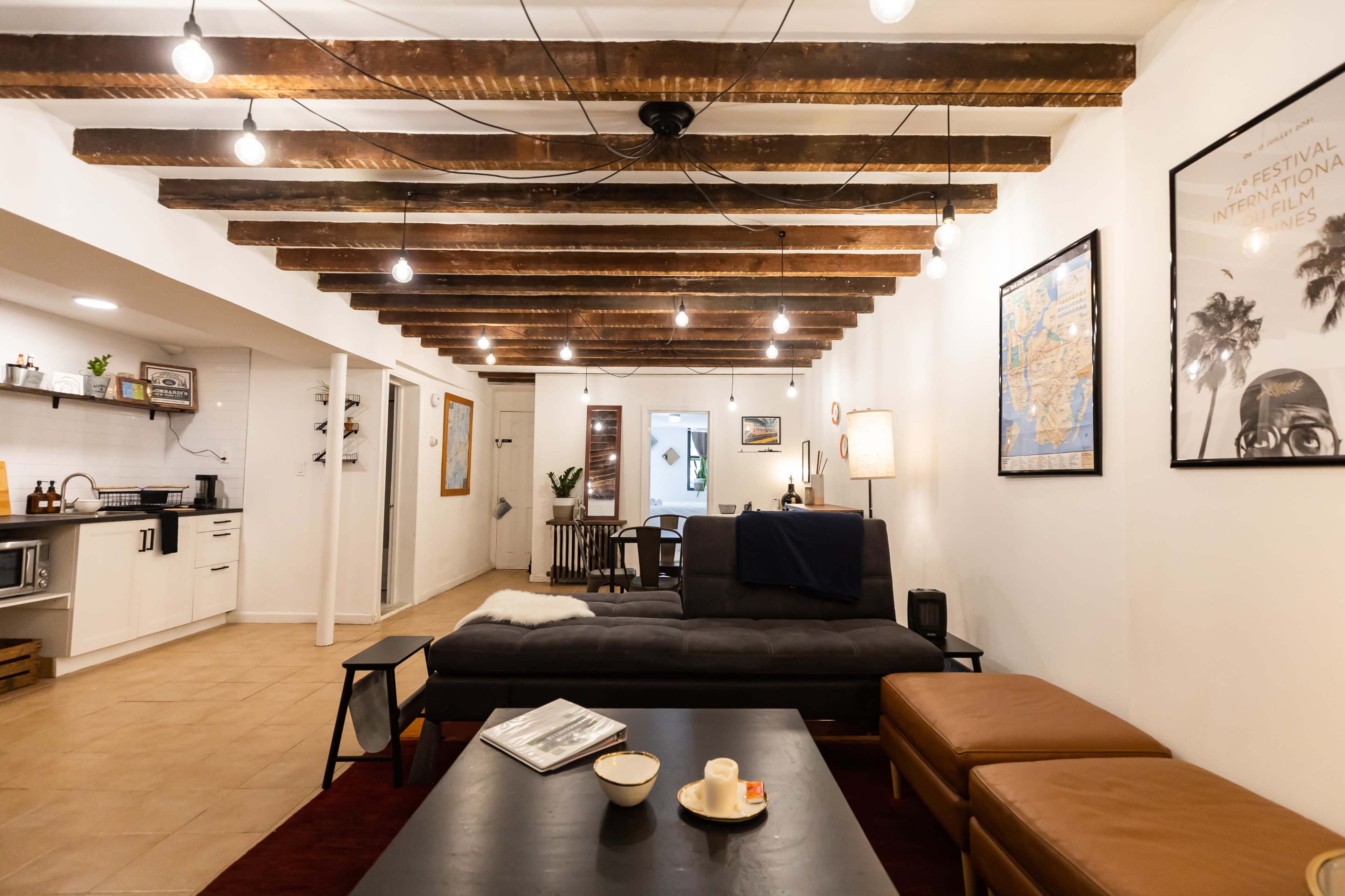 A modern living area with exposed wooden beams, a dark sofa, two brown leather ottomans, and a coffee table on a carpet, illuminated by hanging light bulbs.