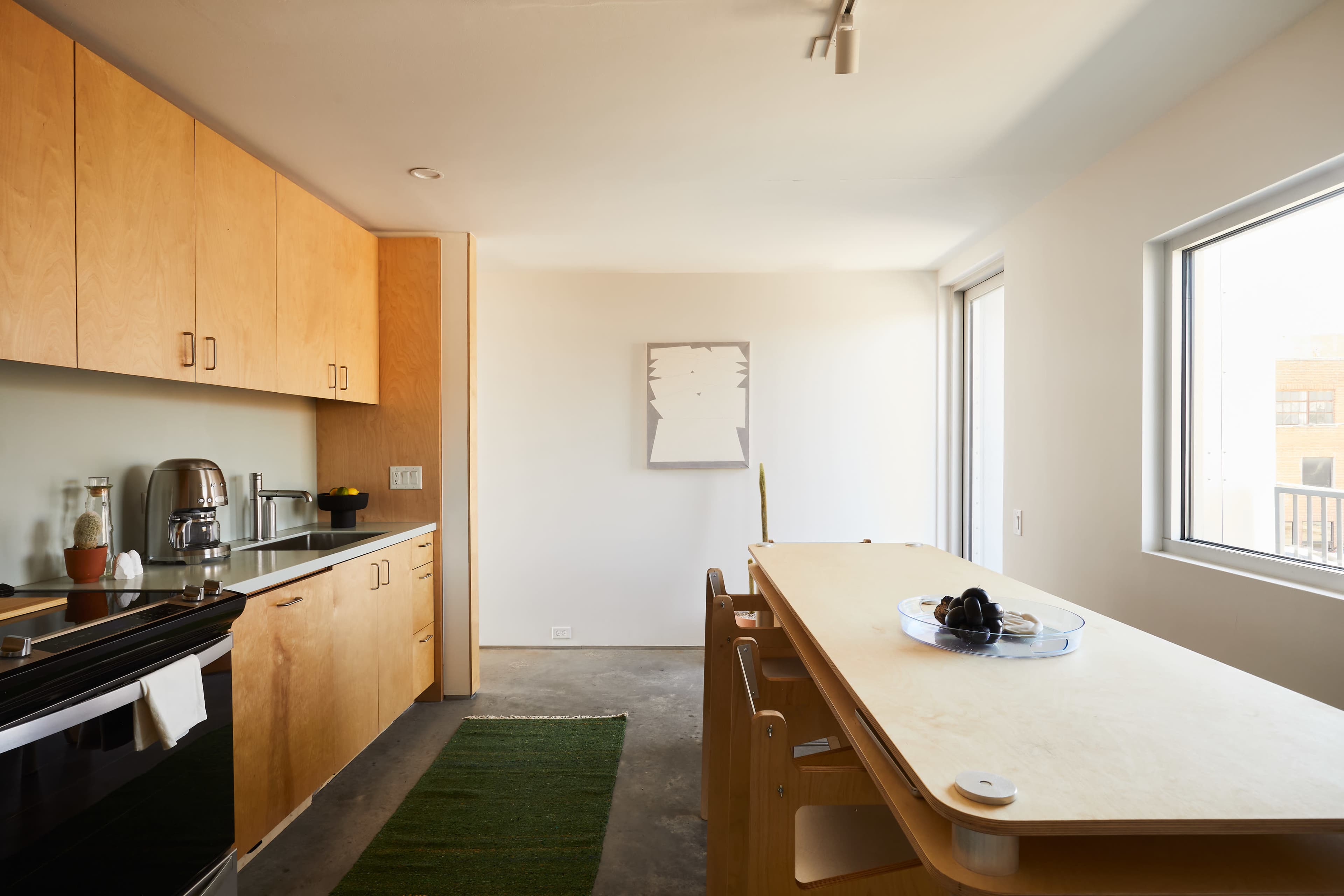 A minimalistic kitchen features wooden cabinetry, stainless steel appliances, and a long table beside large windows.