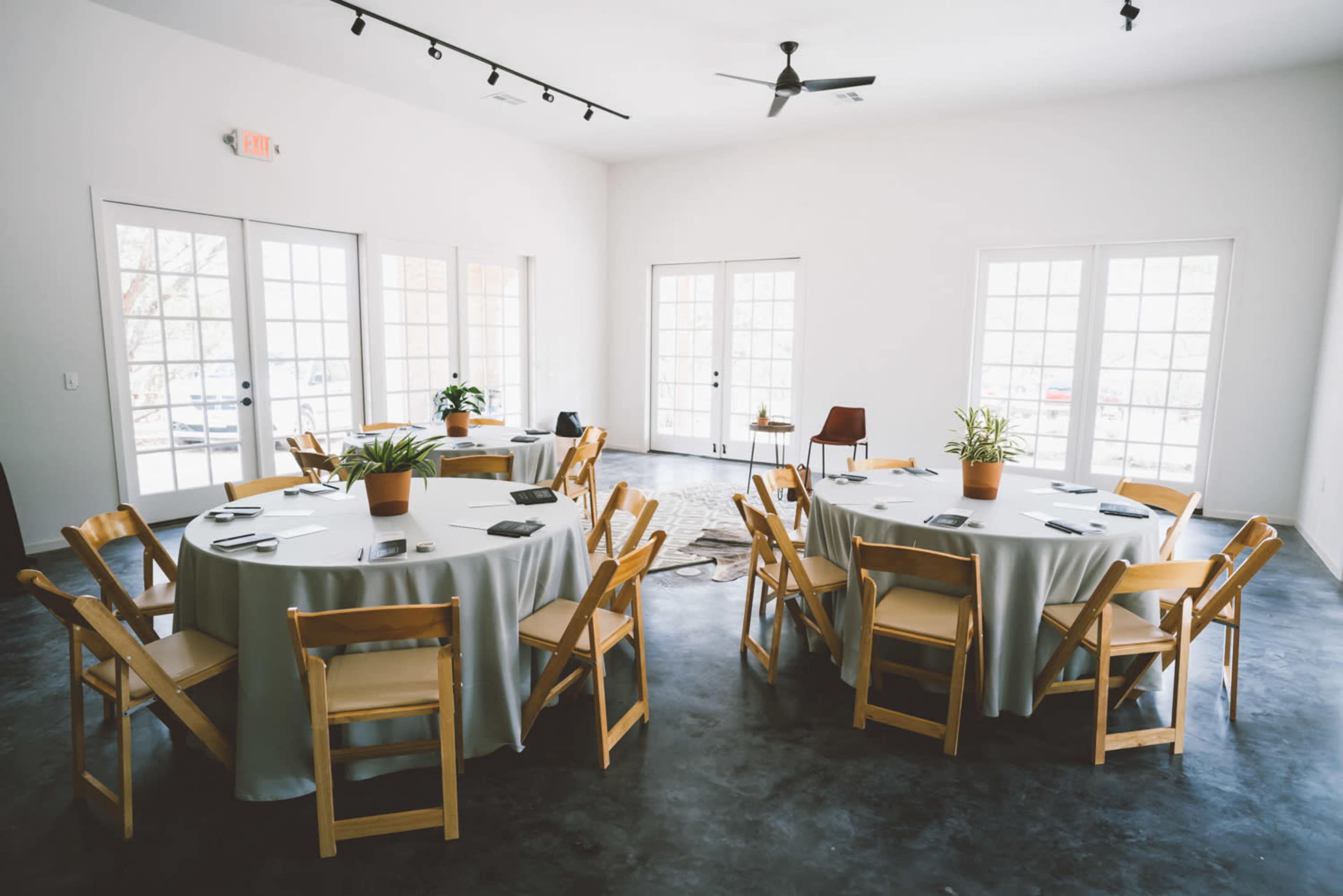 A bright room features multiple round tables with chairs, surrounded by large windows and a concrete floor.