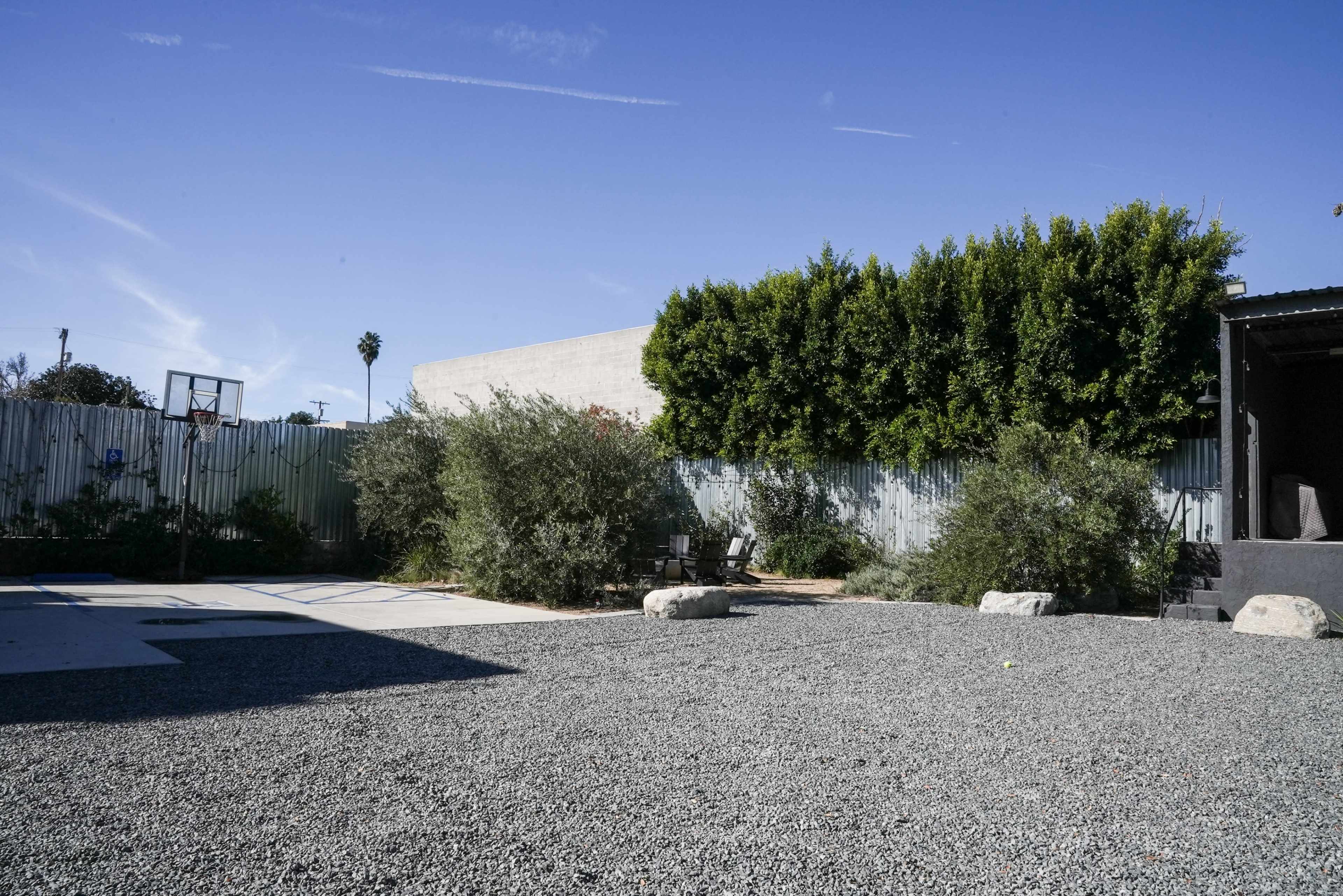 The scene features a gravel yard with scattered rocks, bordered by green shrubs and a wall covered with a fence, under a clear blue sky.