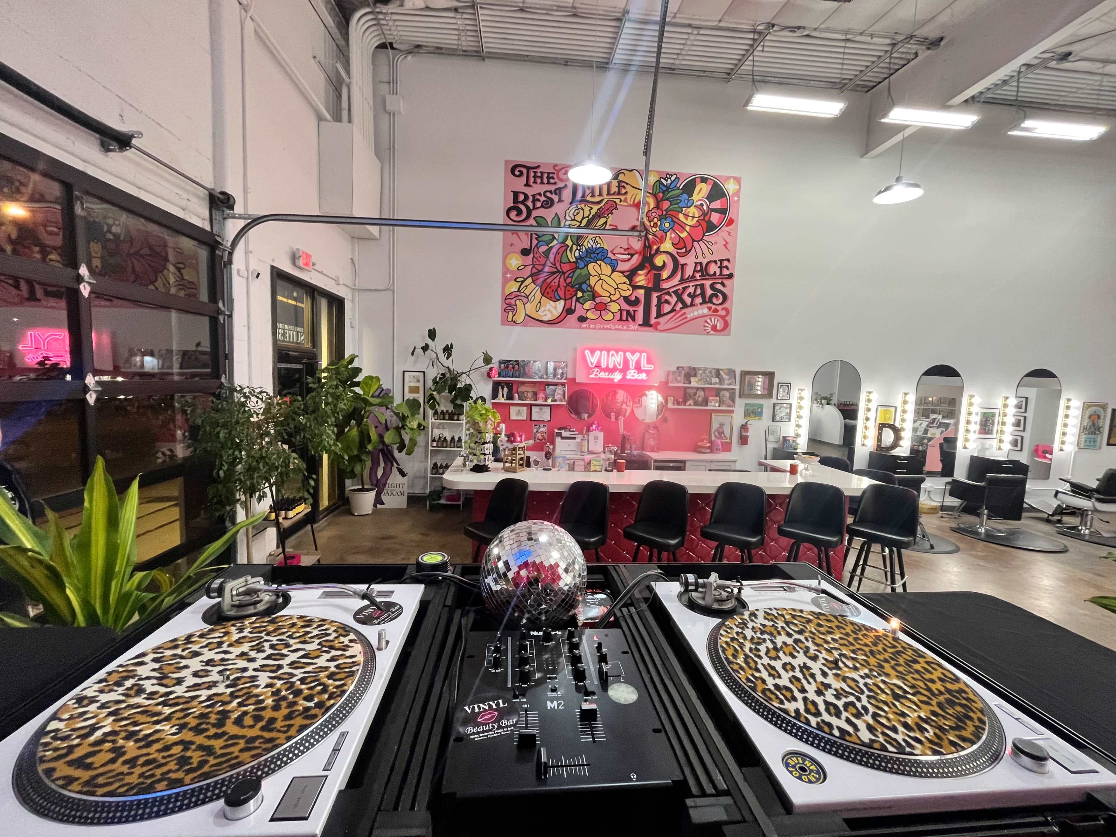 The image shows a vibrant beauty salon featuring a DJ setup with leopard-patterned turntables and a disco ball, surrounded by colorful artwork and salon stations.