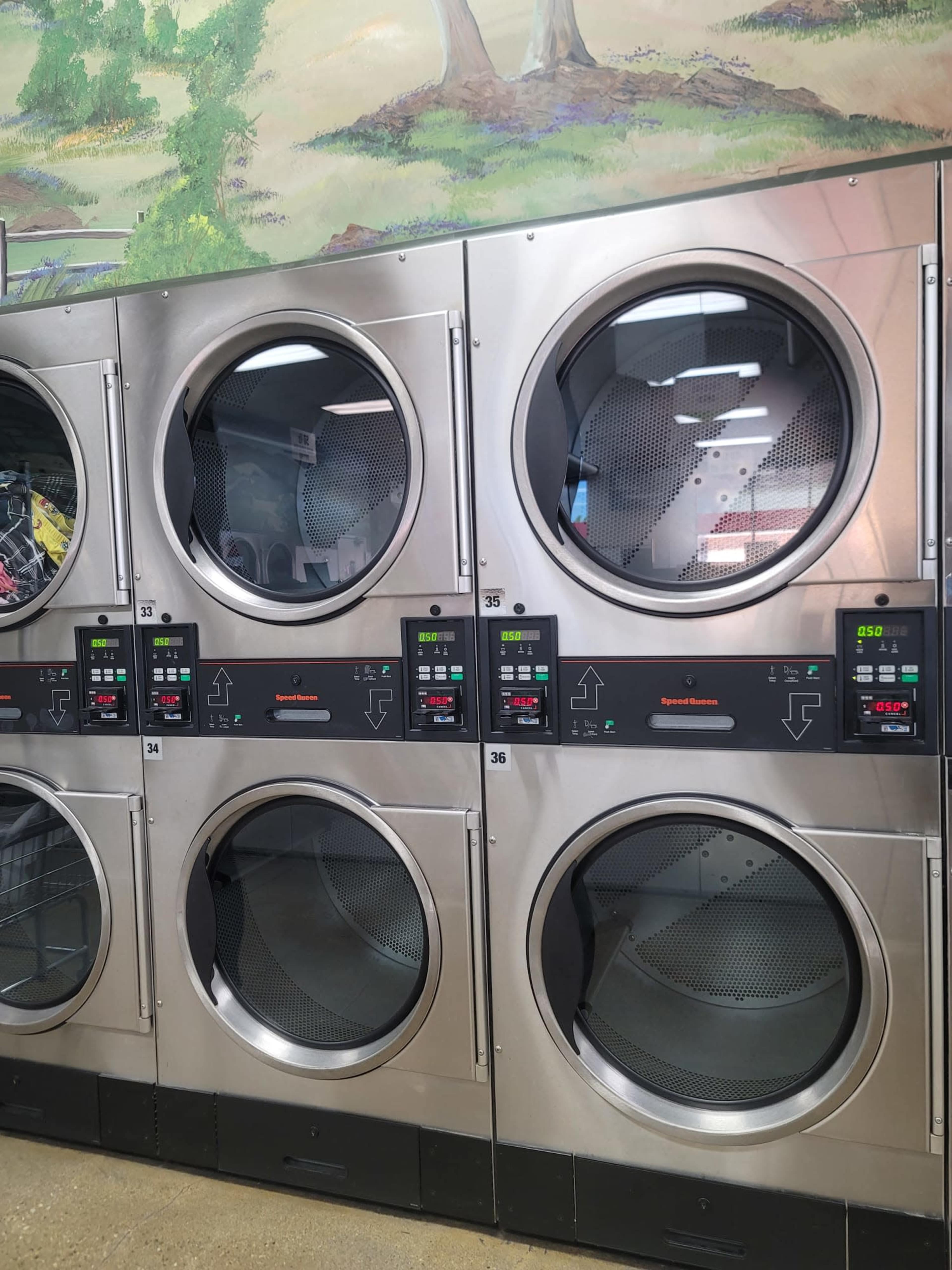 The image shows a row of stainless steel dryers with digital control panels in a laundromat setting.