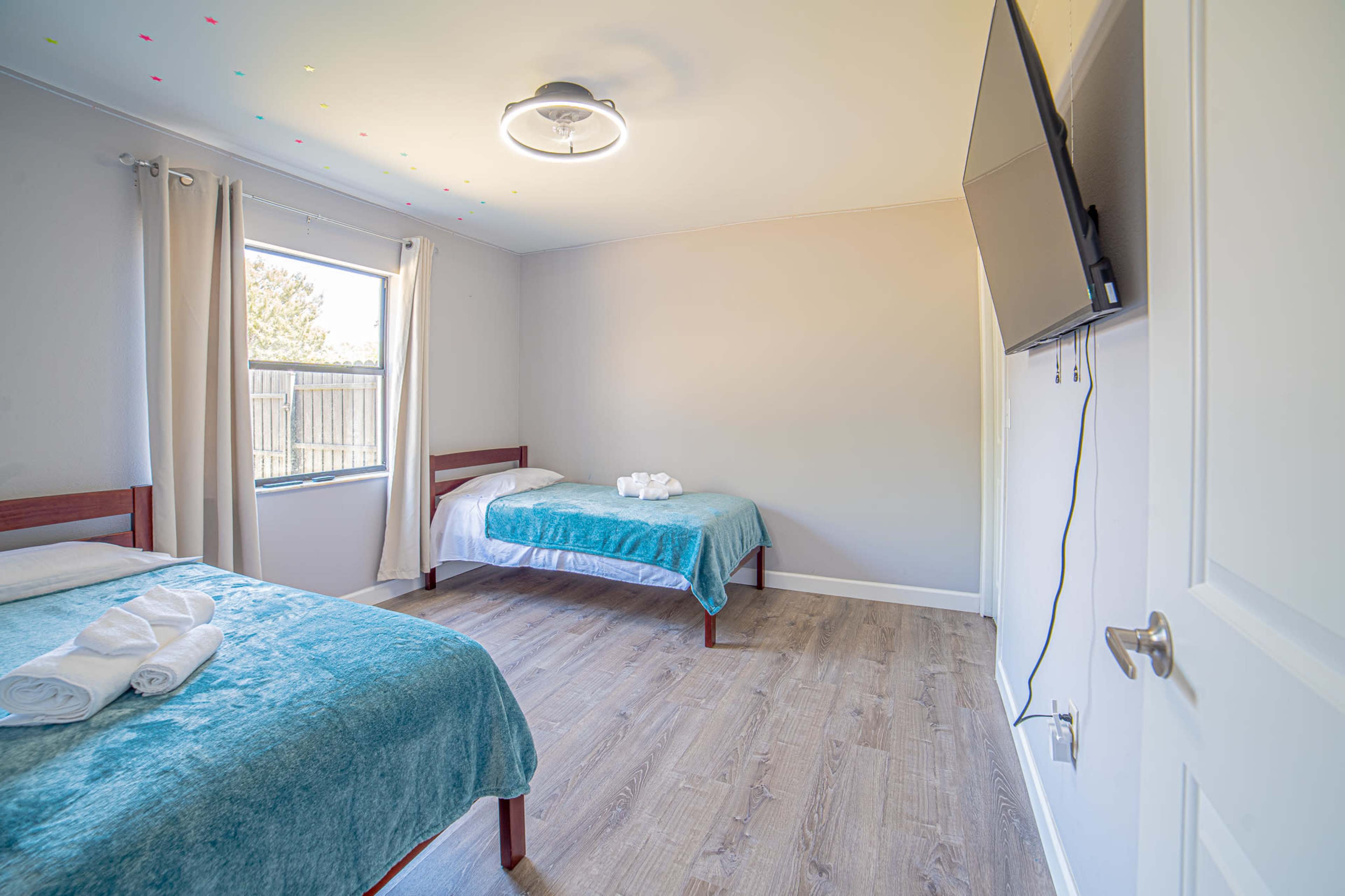 The image shows a simple bedroom with two beds, each covered with teal blankets, a television mounted on the wall, and a window allowing natural light.