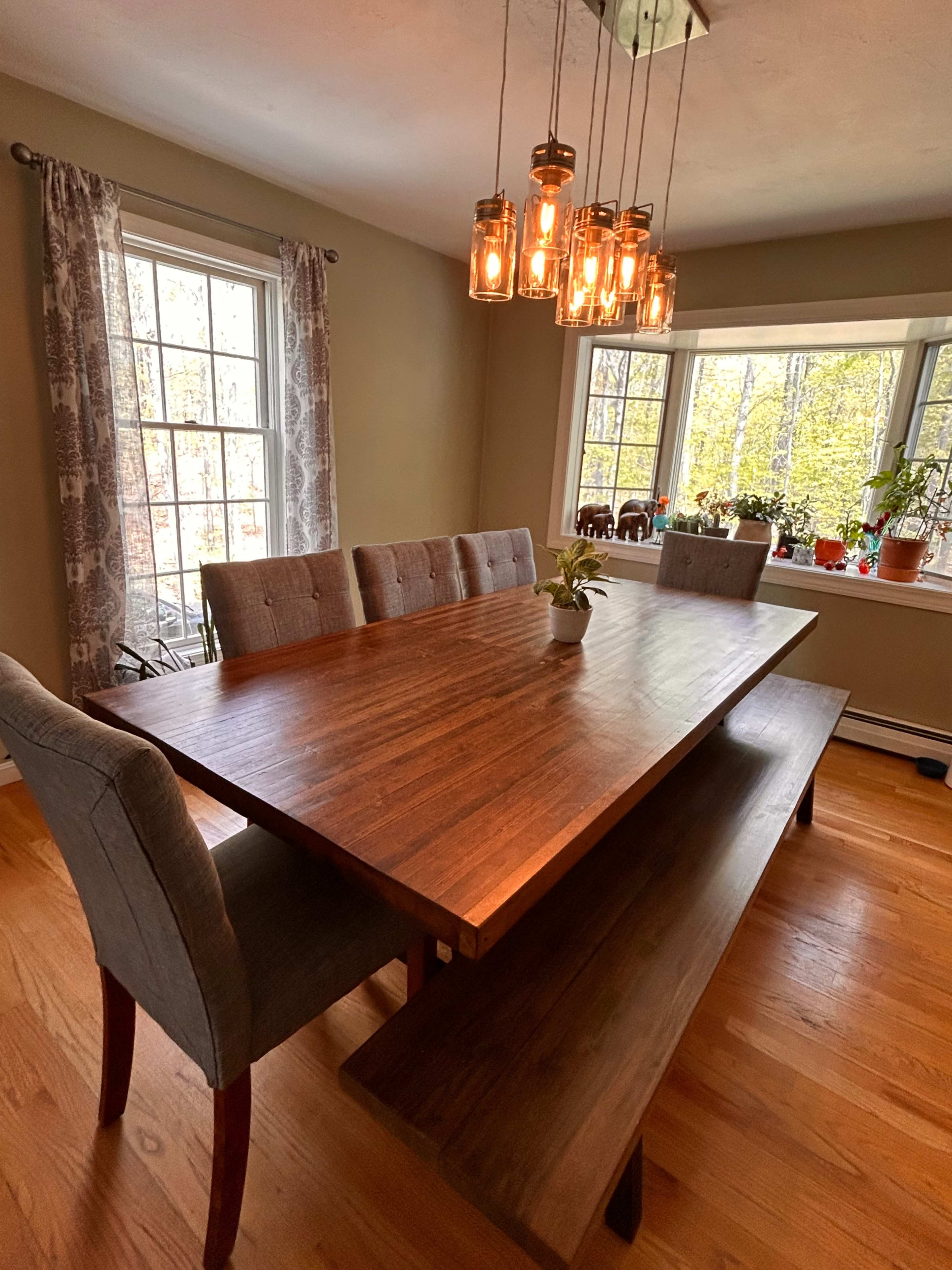 A wooden dining table with a bench and upholstered chairs is set under a cluster of hanging lights in a room with large windows and various plants.