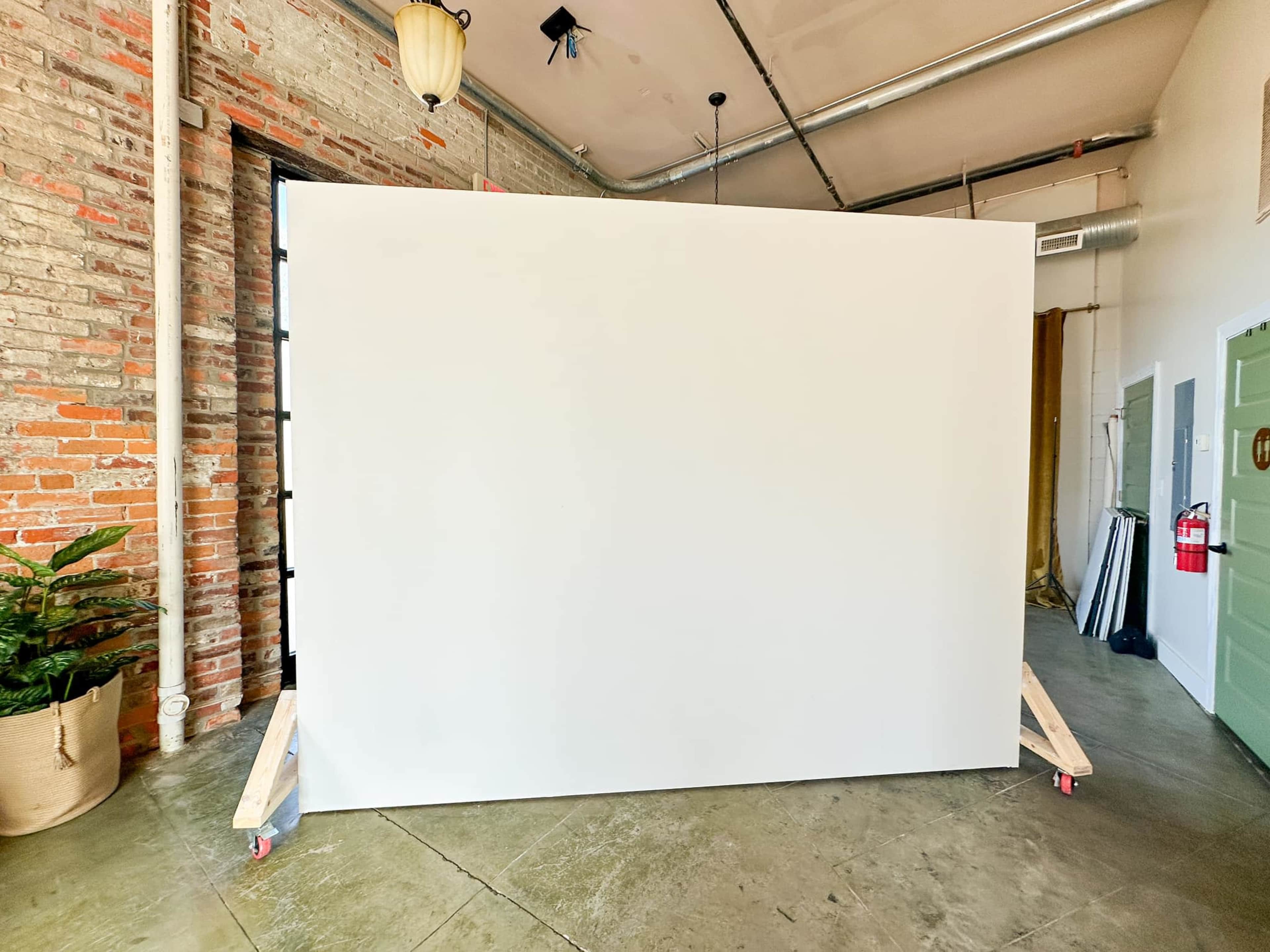 The image shows a large, blank white wall on wheels in an industrial-style room with exposed brick and a concrete floor.