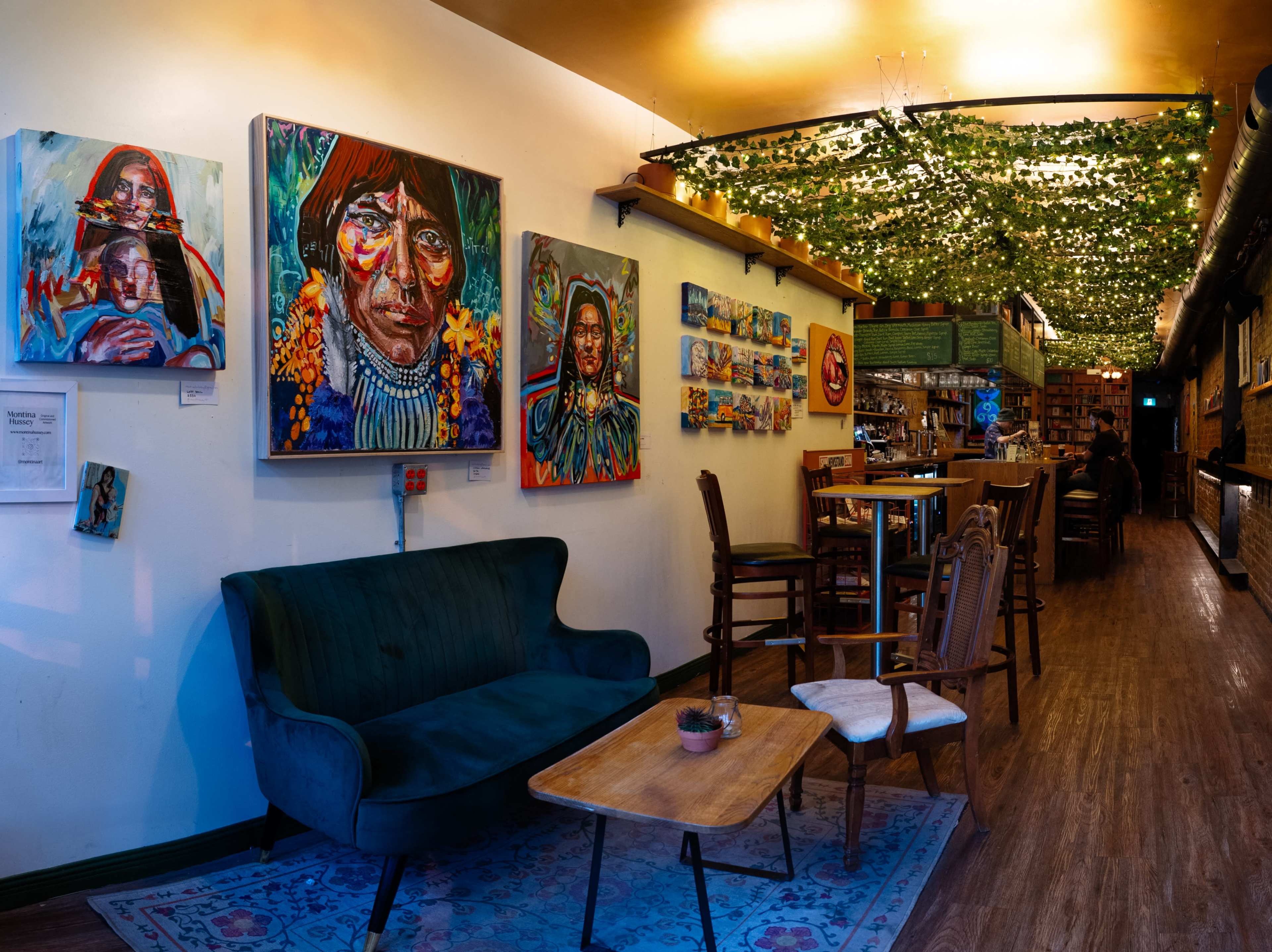 A cozy café interior featuring a green velvet sofa, wooden tables, and vibrant artwork on the walls, with overhead greenery creating a warm atmosphere.