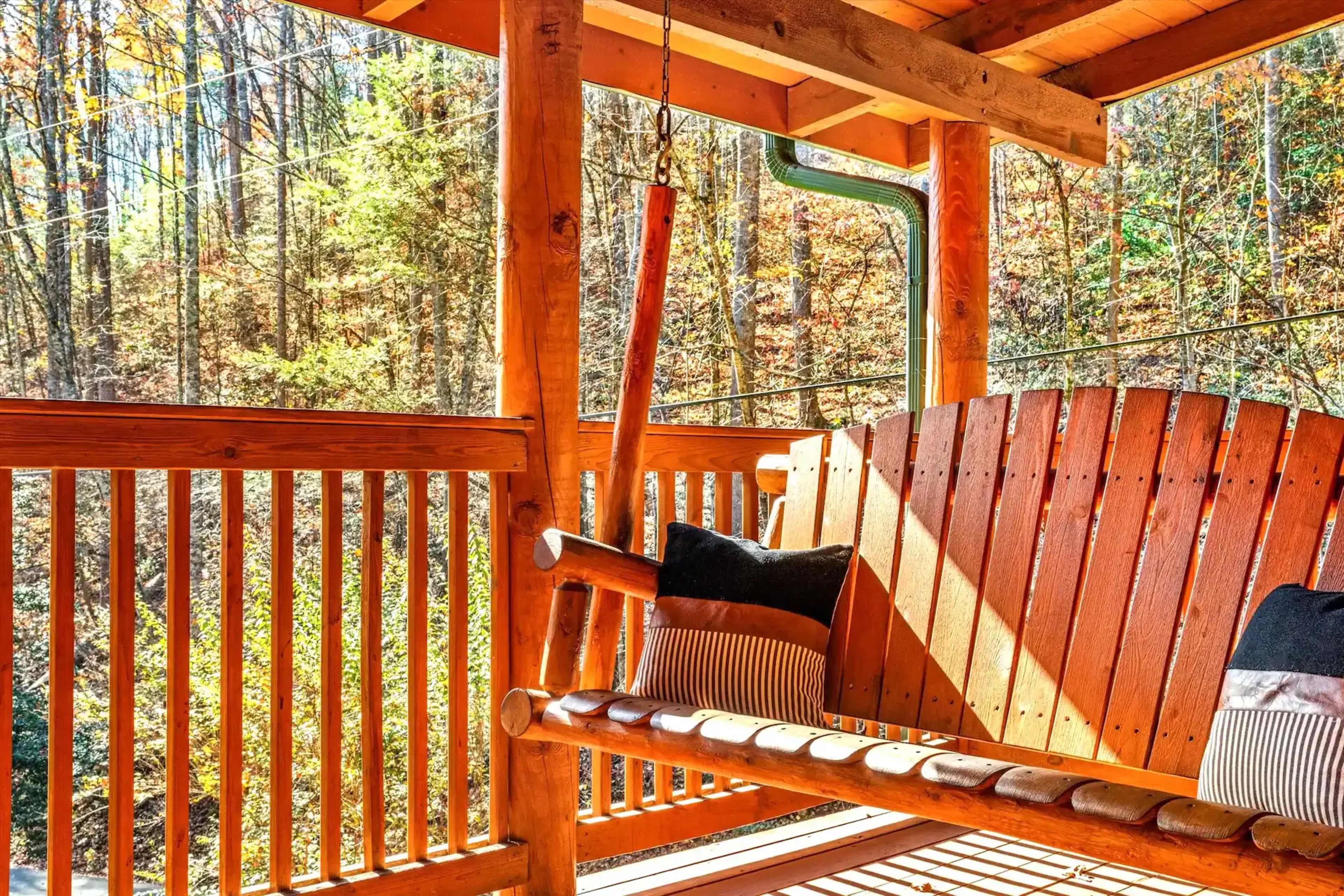 A wooden swing chair with two pillows is positioned on a porch overlooking a forested area with autumn foliage.