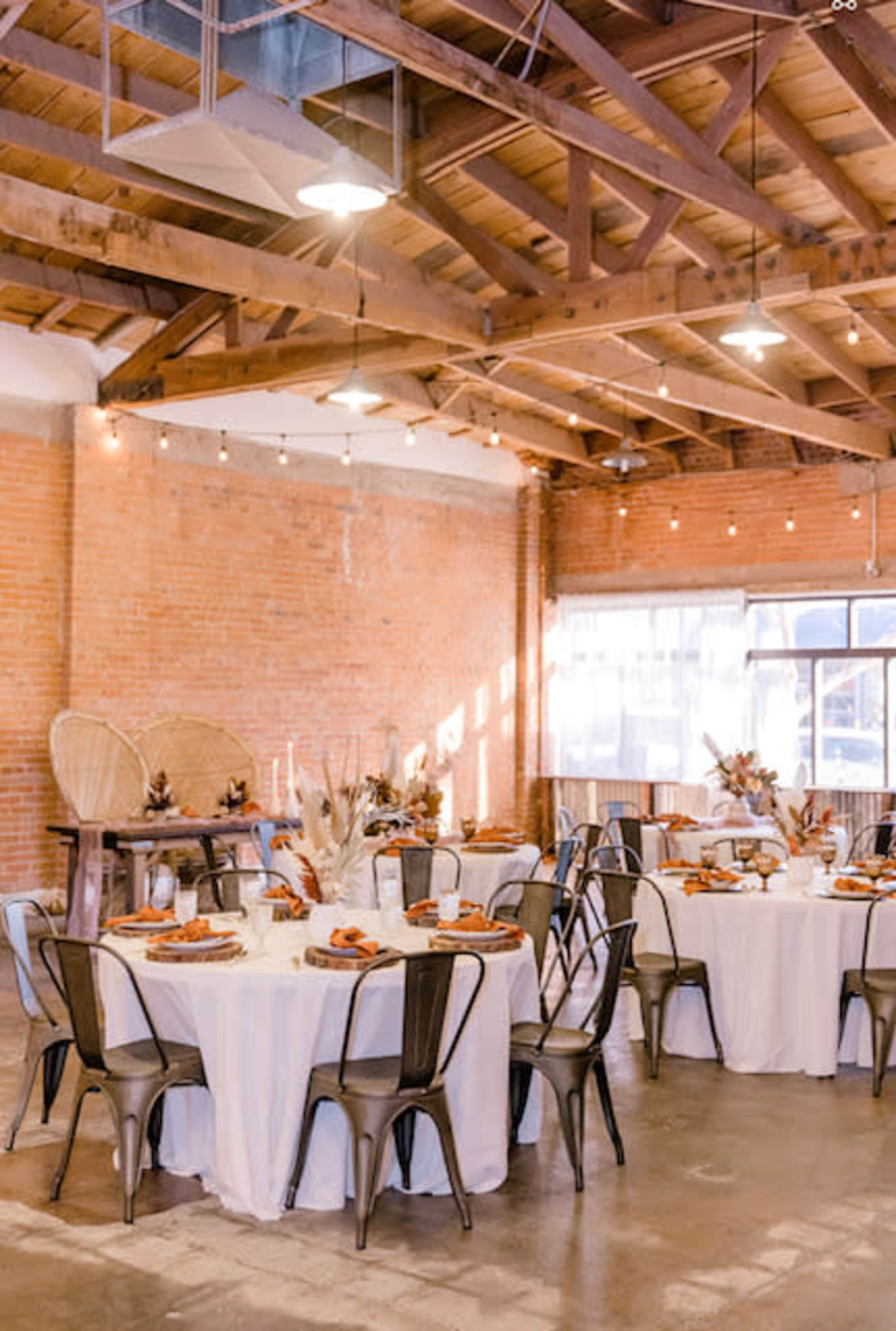 A spacious event venue features round tables with white tablecloths, black metal chairs, and decorative centerpieces, all set against a backdrop of exposed brick walls and wooden beams.