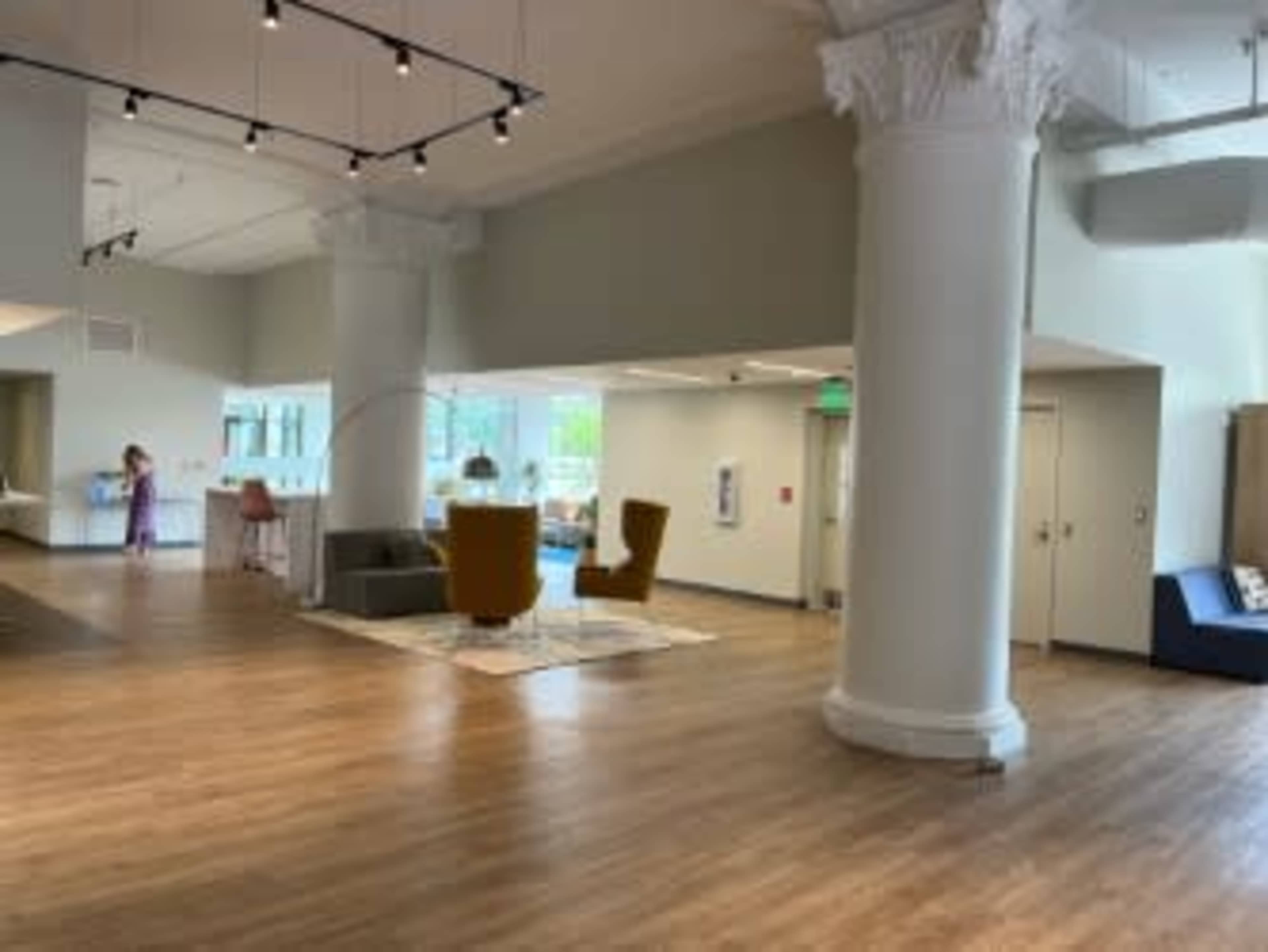 An open interior space featuring wooden flooring, large pillars, a seating area with two chairs, and a person standing in the background.