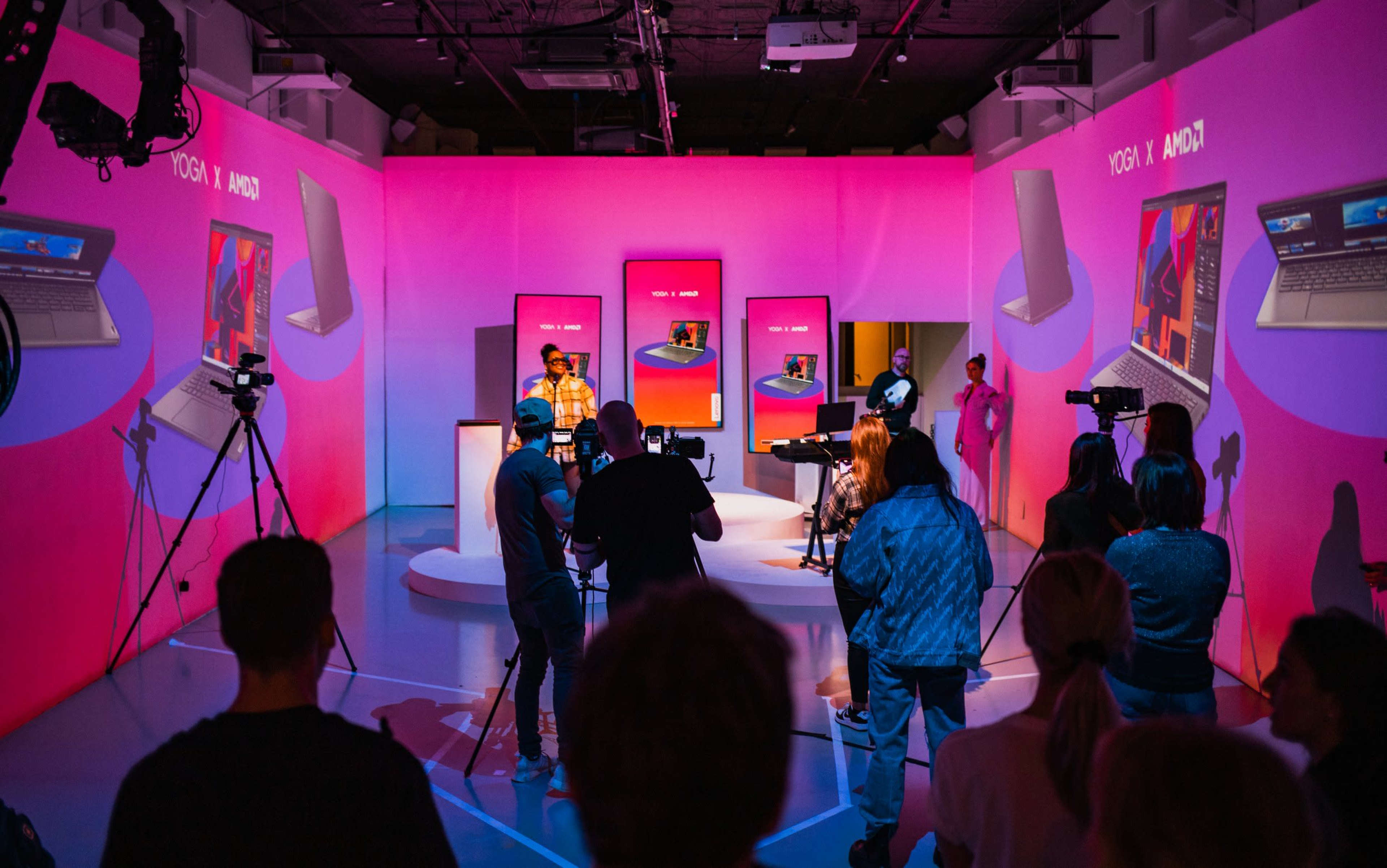 A group of people is gathered in a brightly lit studio with pink backgrounds, filming a promotional event featuring laptops and presentations.