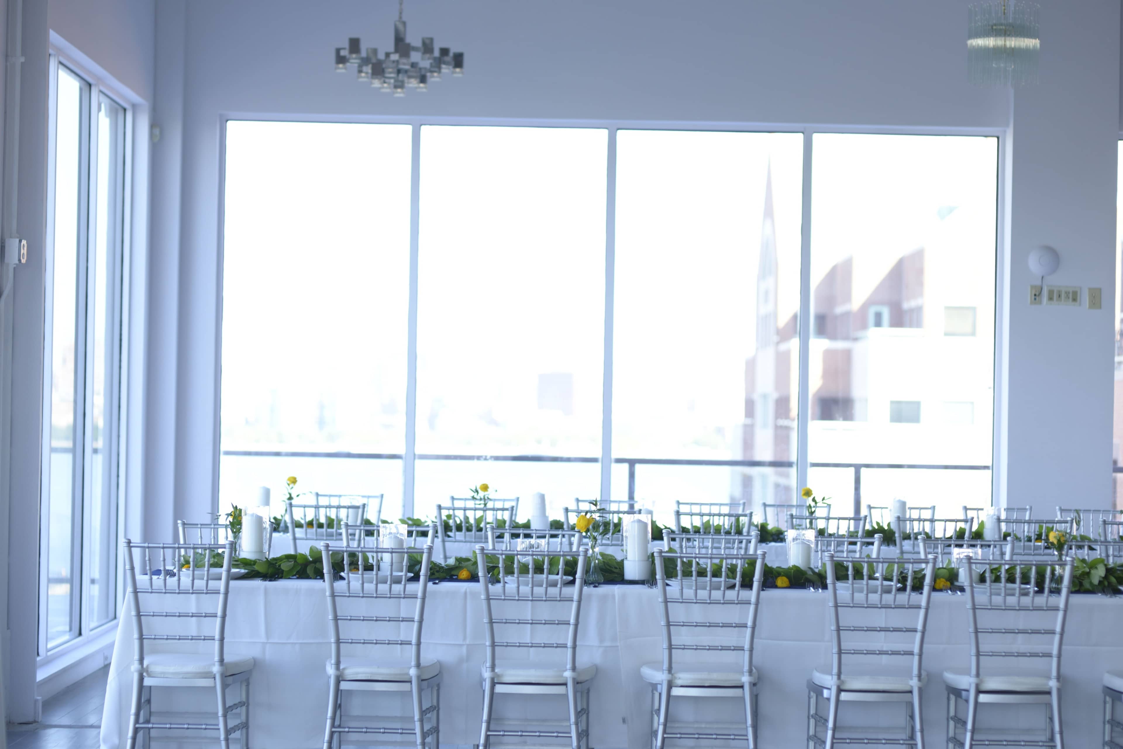 A banquet hall features a long, elegantly set table adorned with greenery and candles, positioned near large windows that provide ample natural light.