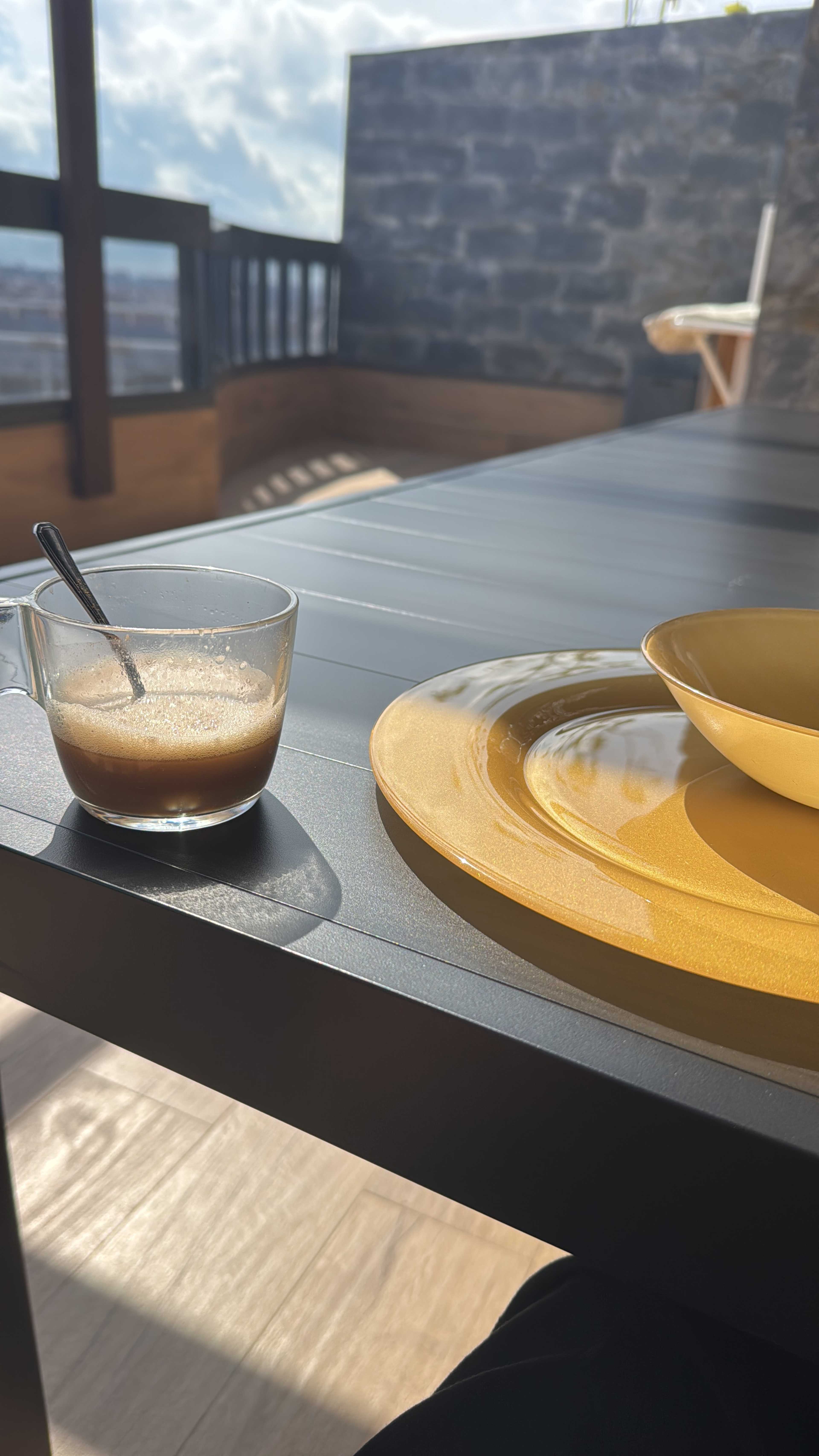 A glass of coffee sits beside a yellow plate and bowl on a table with a mountainous backdrop and a cloudy sky.