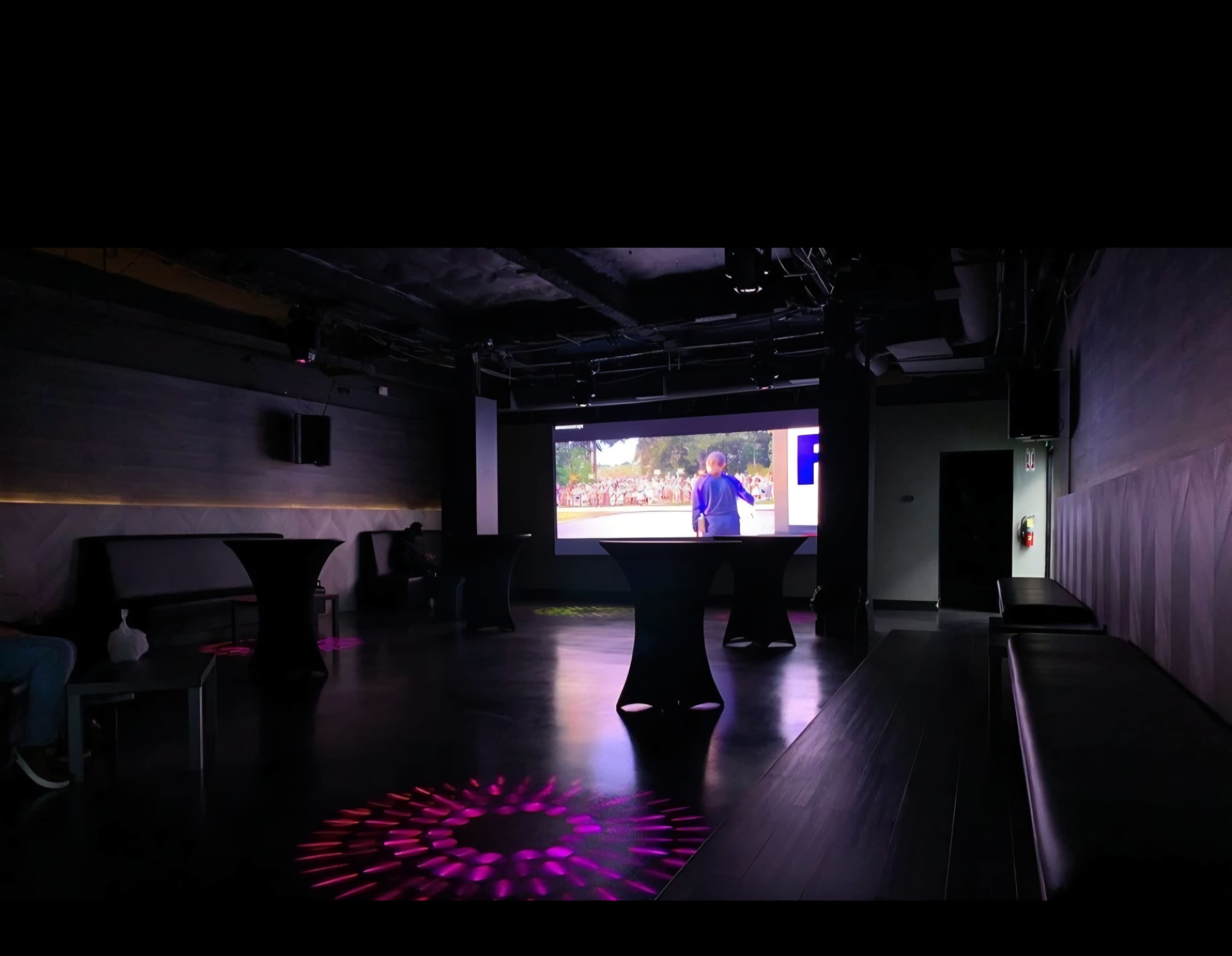 The image shows a dimly lit venue featuring a large screen projecting a video, with scattered black tables and colorful patterns on the floor.