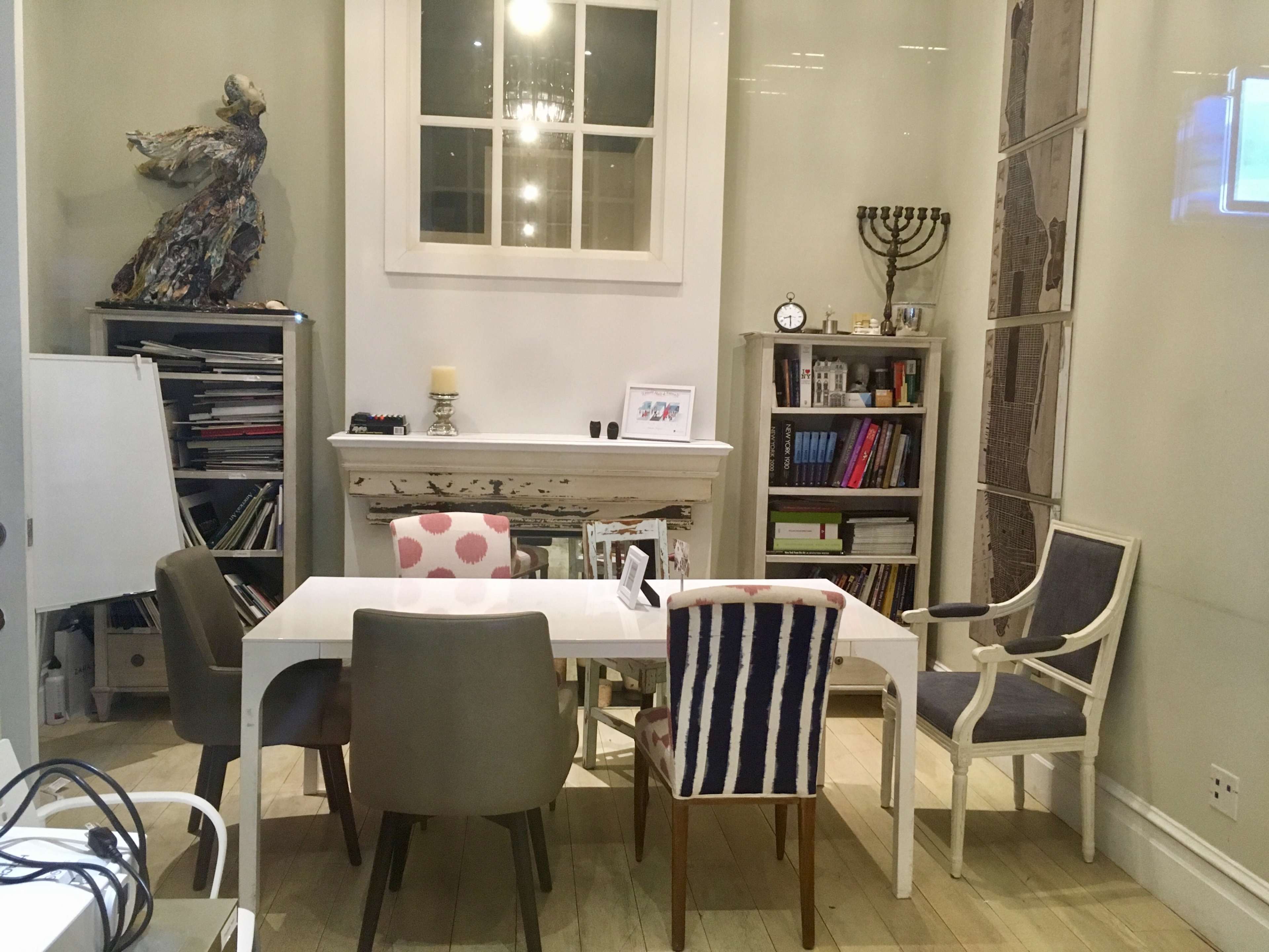A modern meeting room features a white table surrounded by four chairs and a bookshelf against the wall, with art displayed on shelves and a large window.