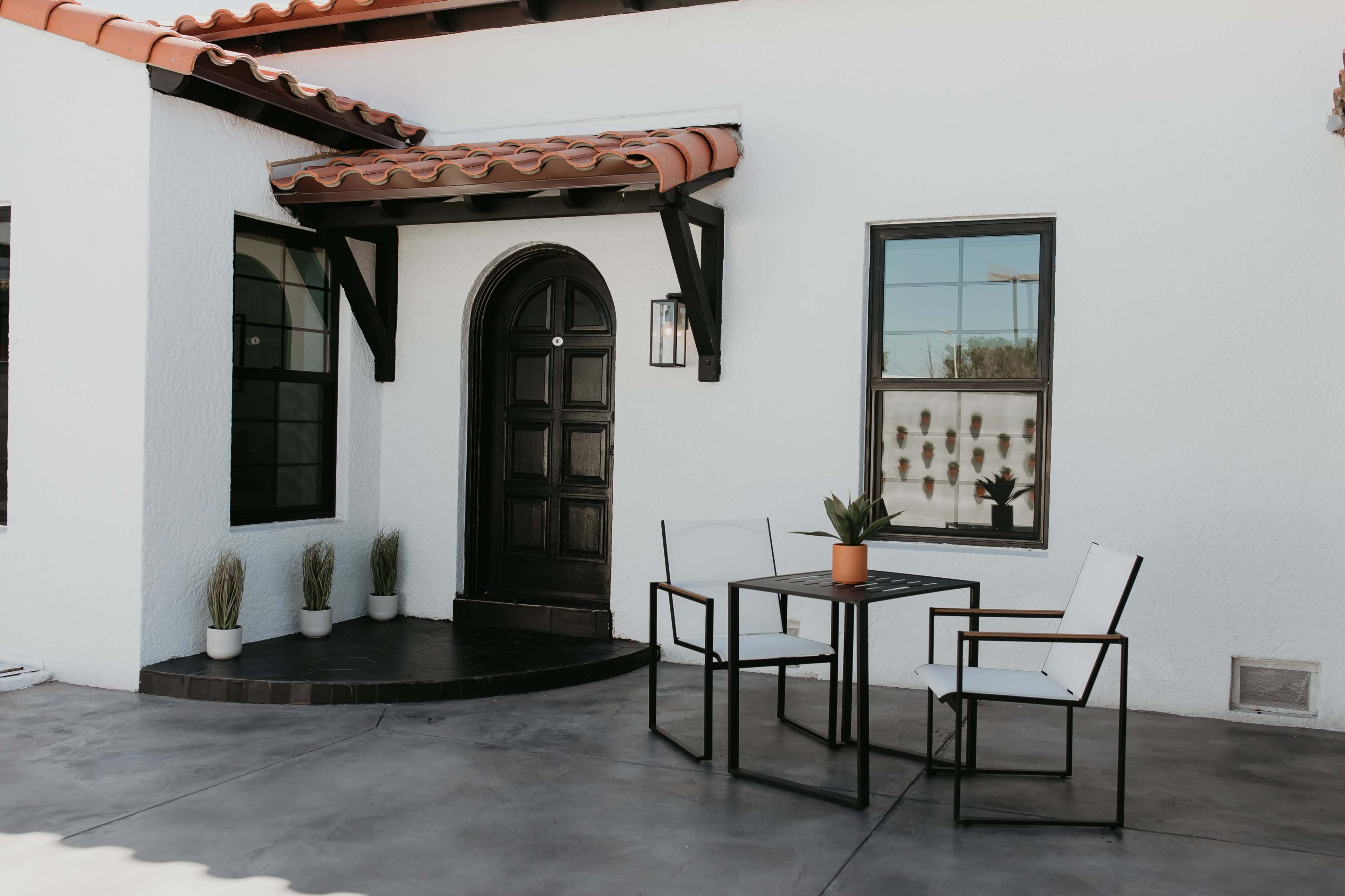 A small outdoor seating area with two chairs and a table is situated in front of a white house with a black door and decorative window.