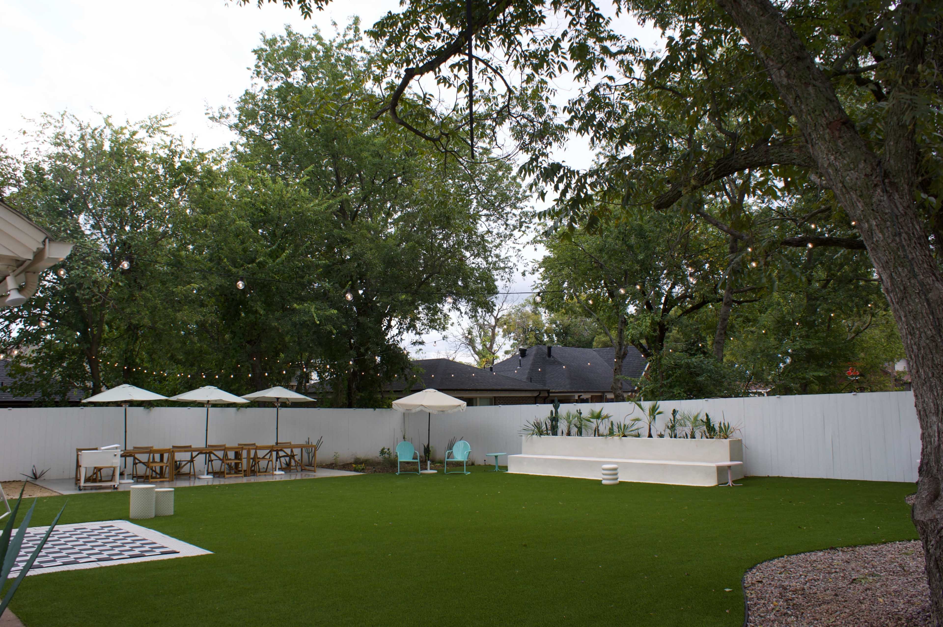The image shows a spacious backyard featuring artificial grass, patio furniture, a dining area under an umbrella, and a white wall with greenery in planters.