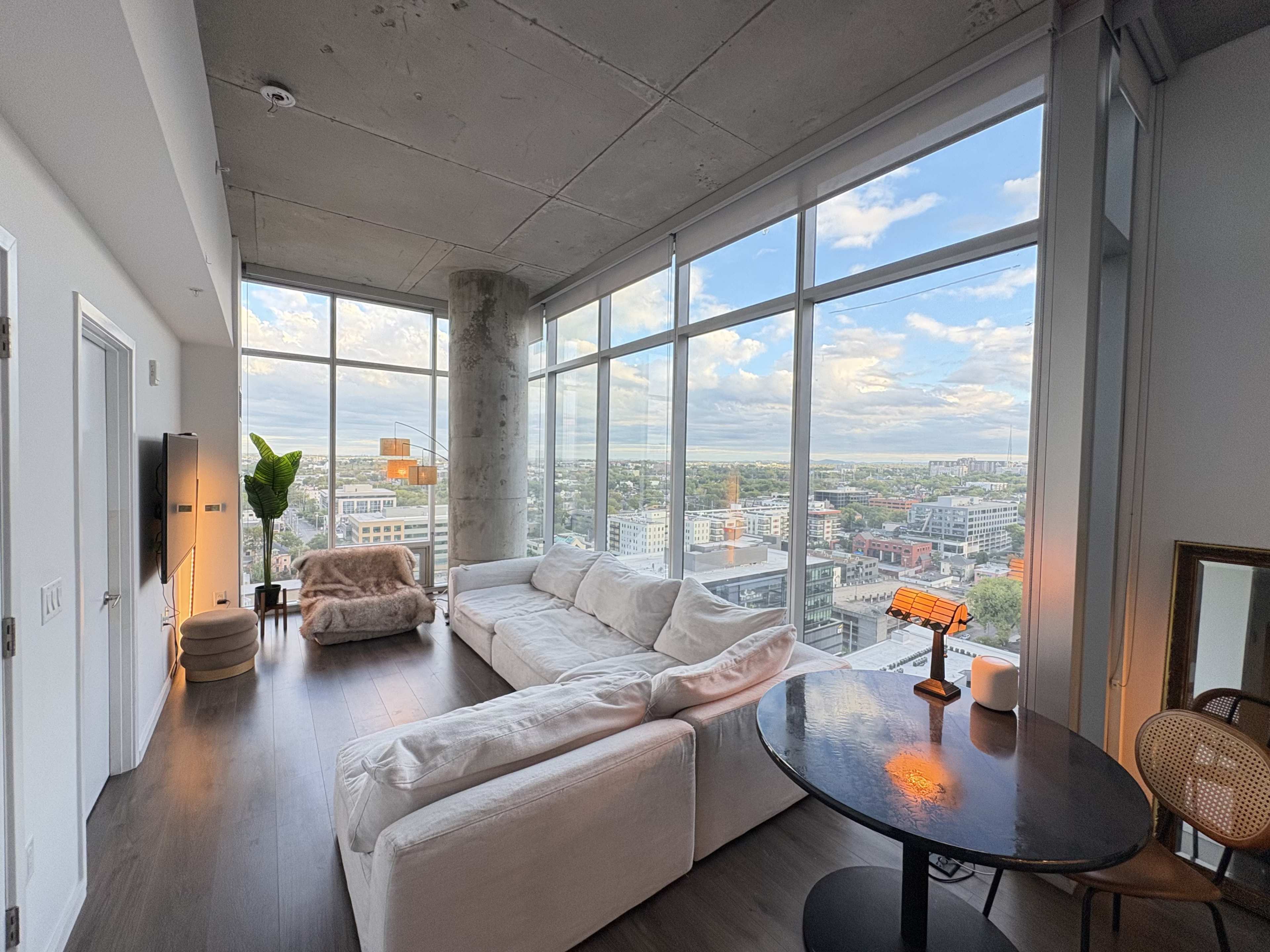 A modern living room features large windows with a view of the city, a white sectional sofa, a small round table, and decorative plants.