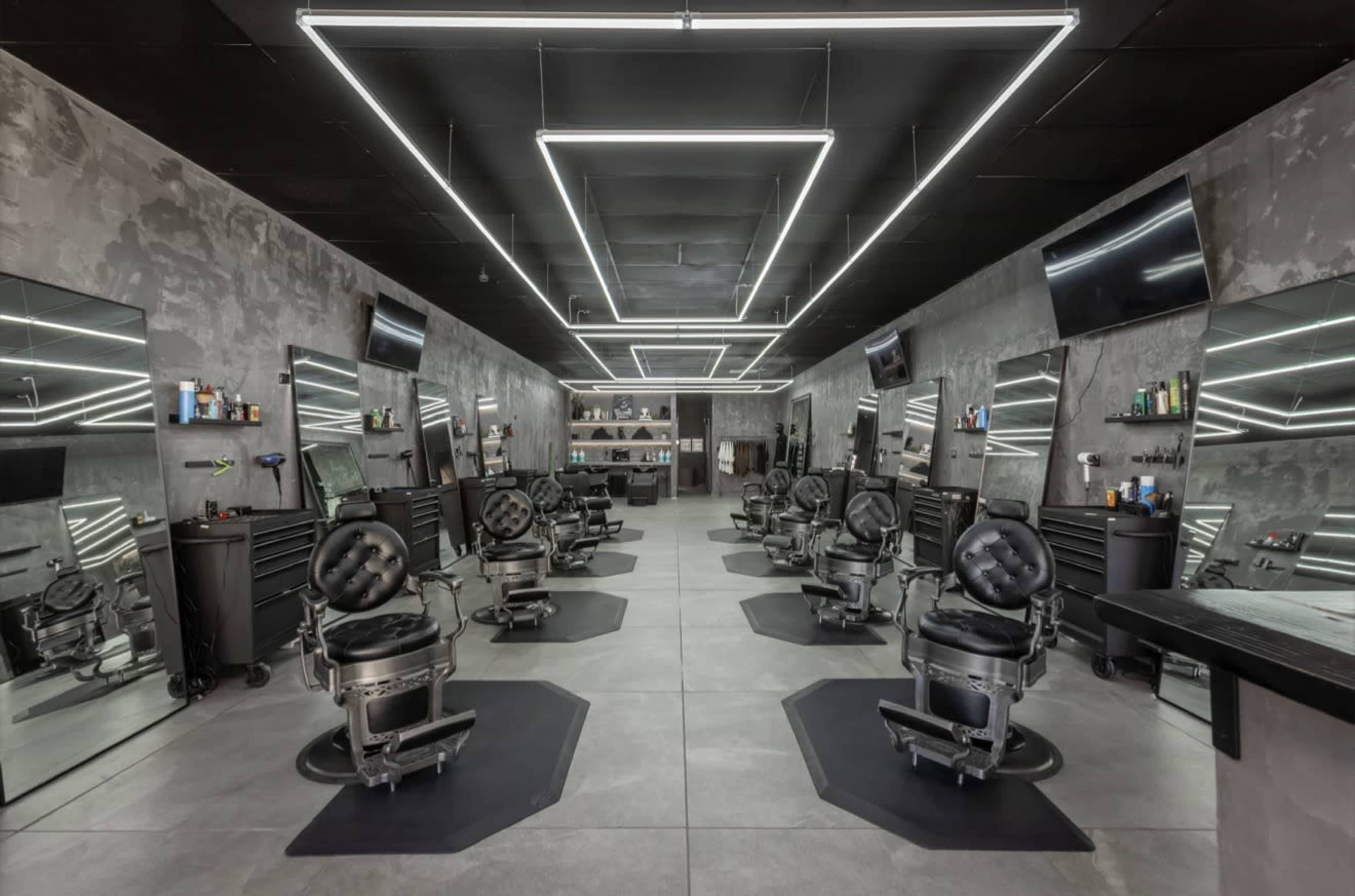 The image shows a modern barber shop with several black barber chairs arranged in a spacious, well-lit interior featuring mirrored walls and sleek lighting.