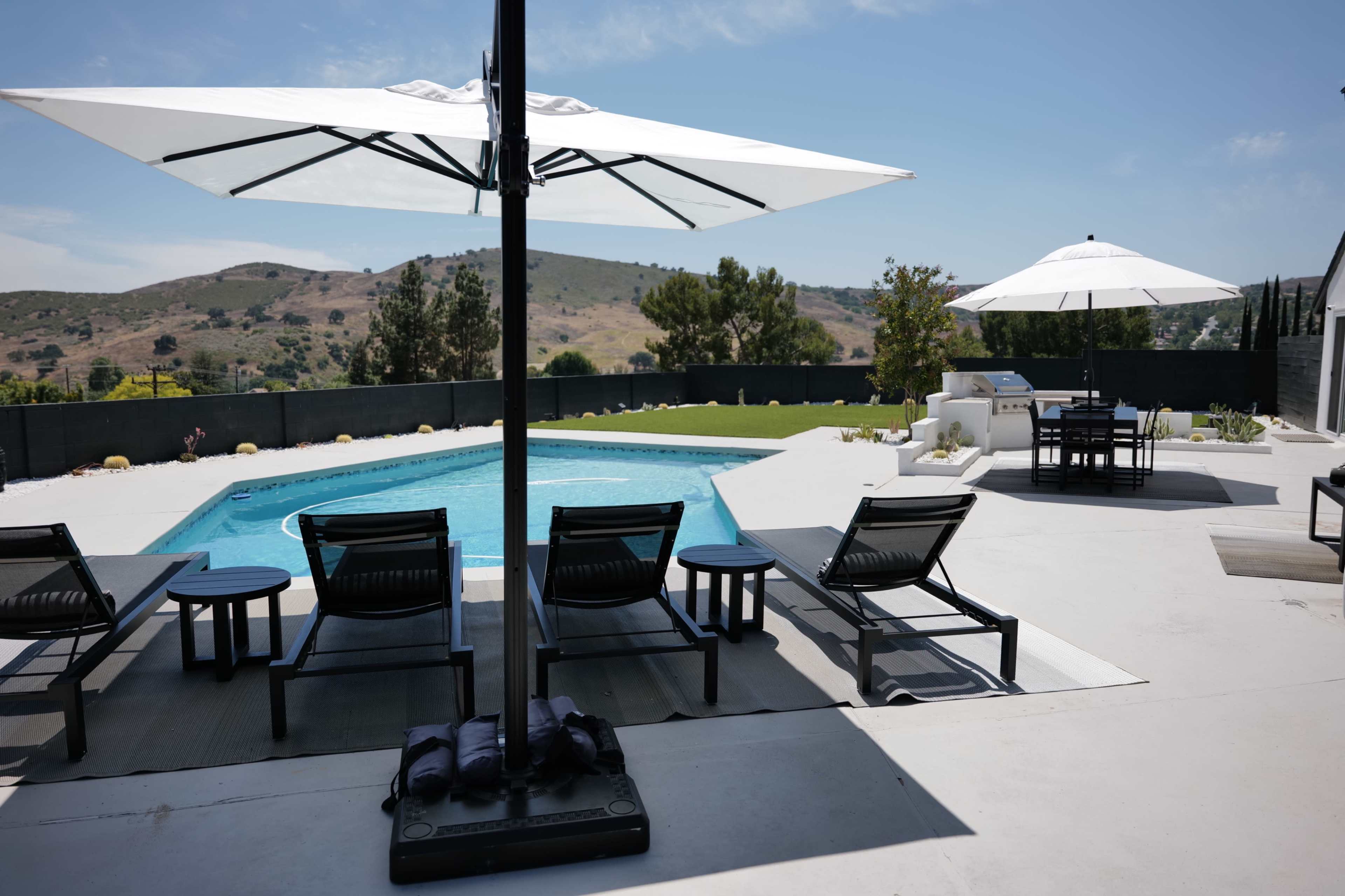 The image shows a modern outdoor area featuring a swimming pool surrounded by lounge chairs and umbrellas, with a landscaped hillside in the background.