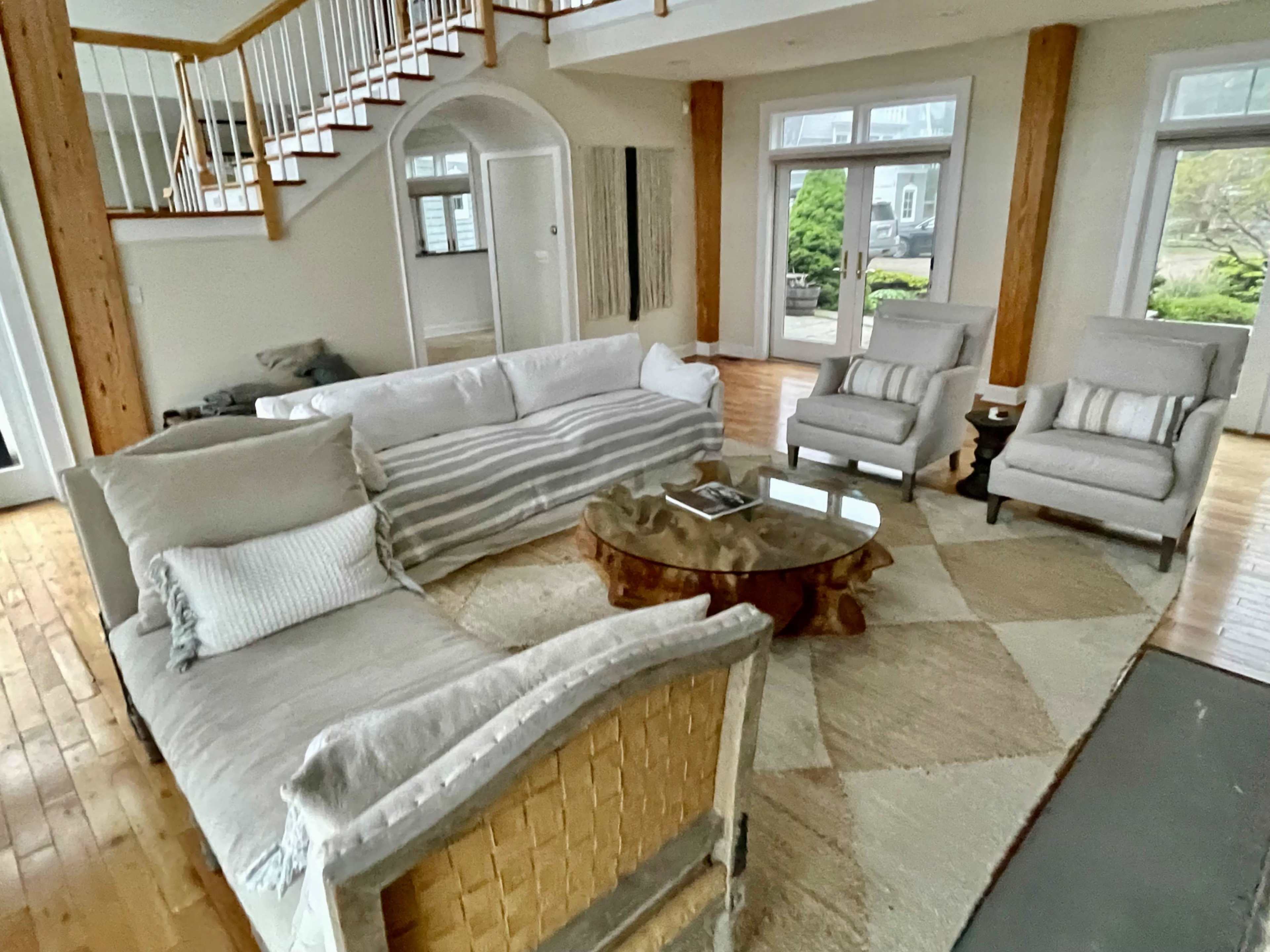 A spacious living room features a sectional sofa, two armchairs, a round wooden coffee table, and large windows overlooking a garden.