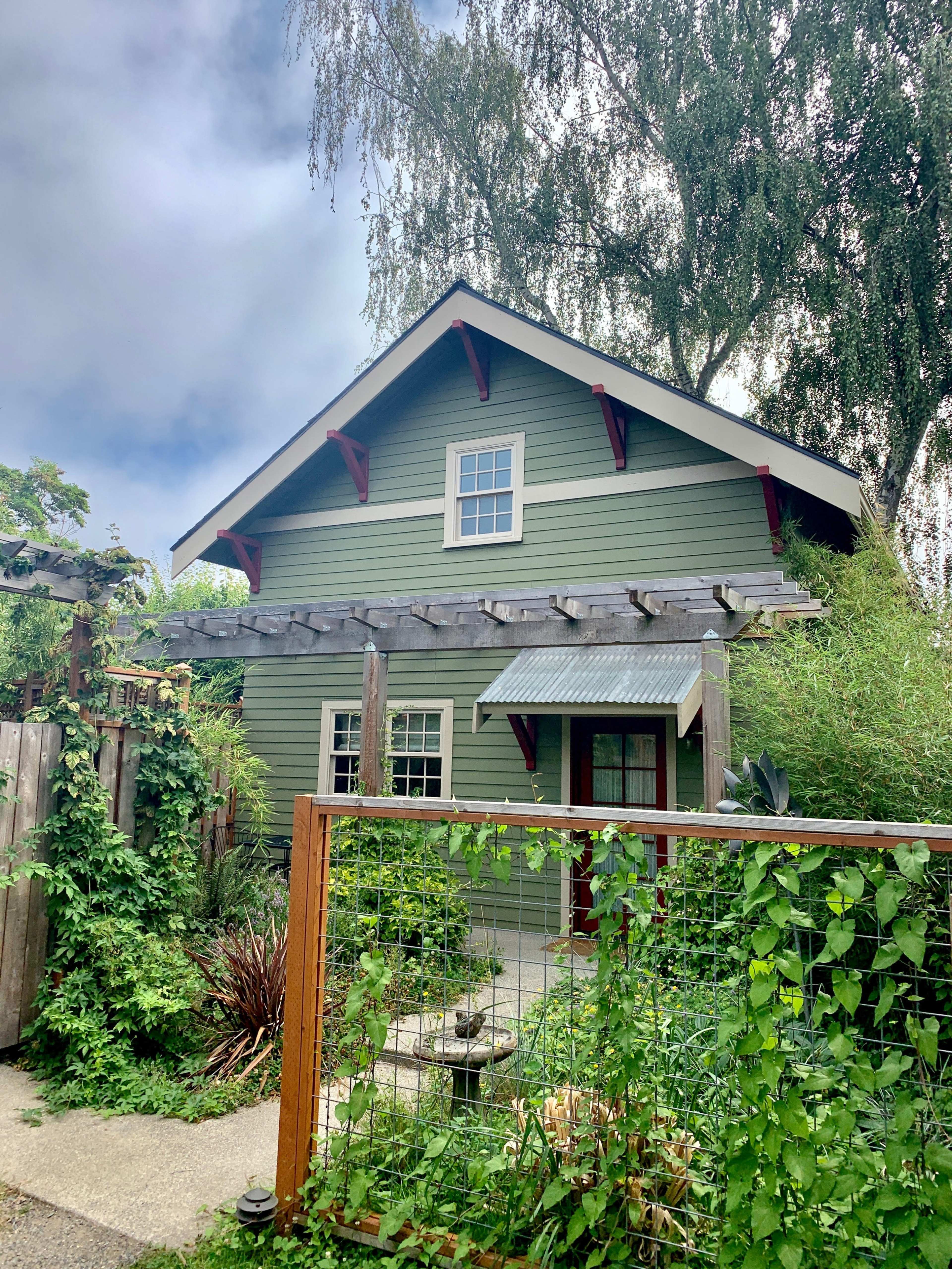 A green house with a white roof and red accents is partially obscured by greenery and a wooden fence.