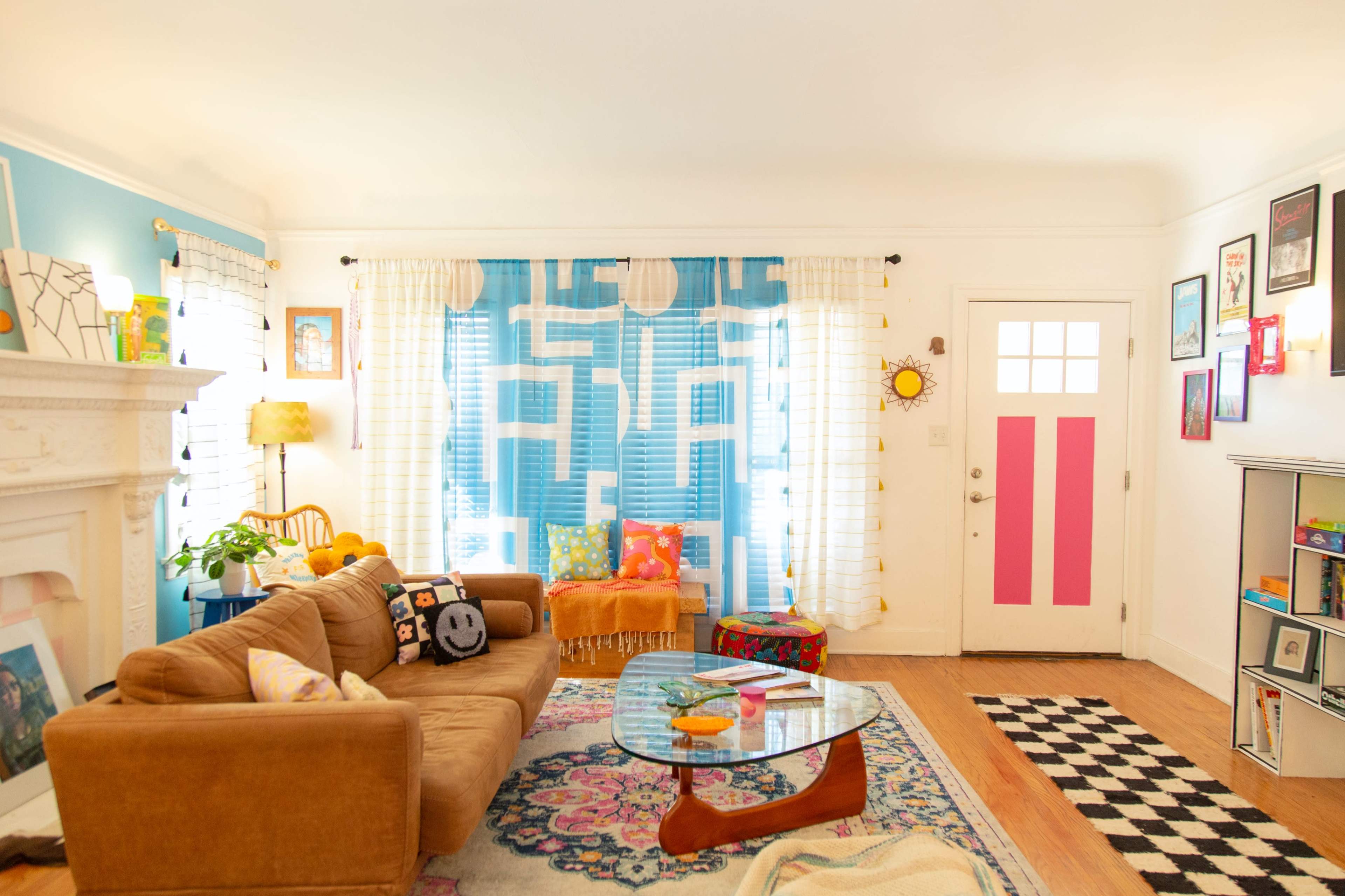 A cozy living room features a brown sofa, a glass coffee table, colorful pillows, and bright blue curtains, with a pink door accentuating the space.