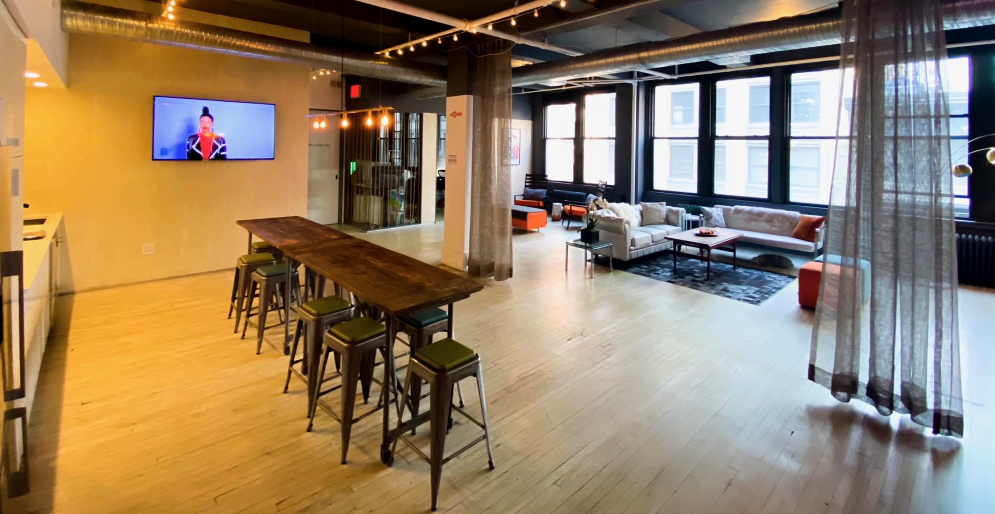 A spacious, modern open-concept room featuring a long wooden table with bar stools, a seating area with a sofa, and a television mounted on the wall.