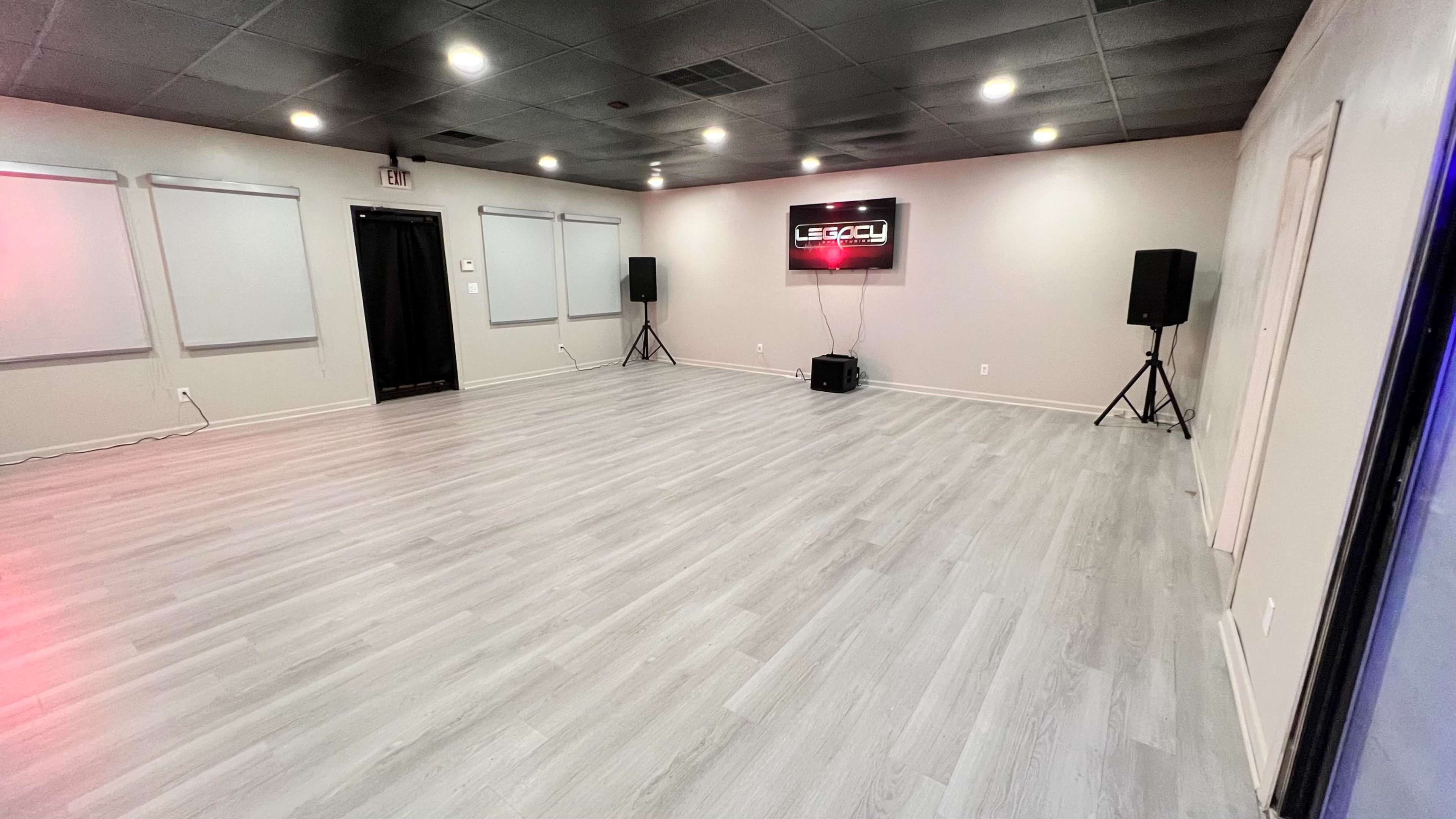 The image shows an empty studio space with gray flooring, wall-mounted screens, and two speaker stands in the corners.