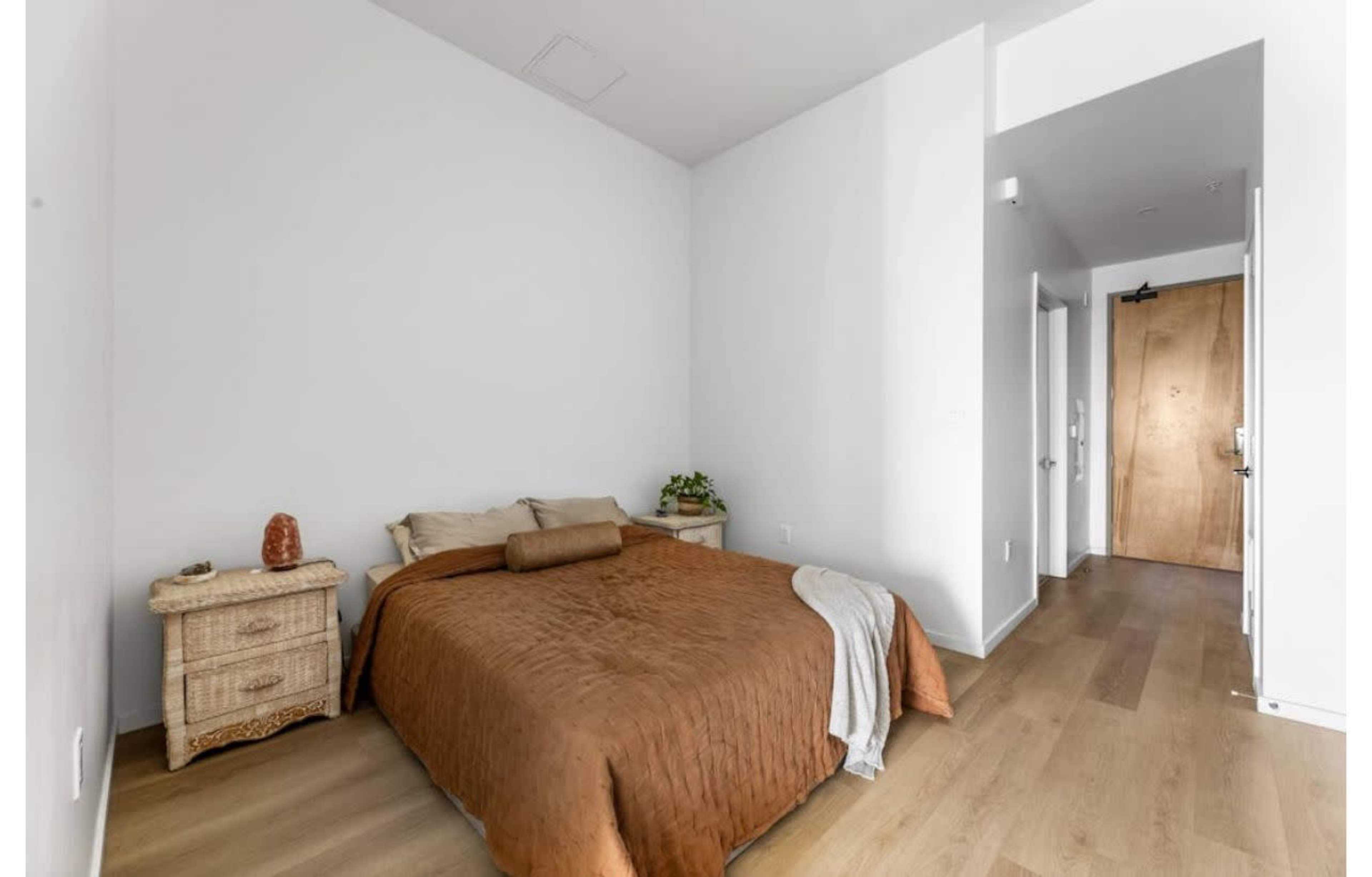 A simple bedroom features a brown quilted bedspread, a bedside table, and a door leading to a hallway.