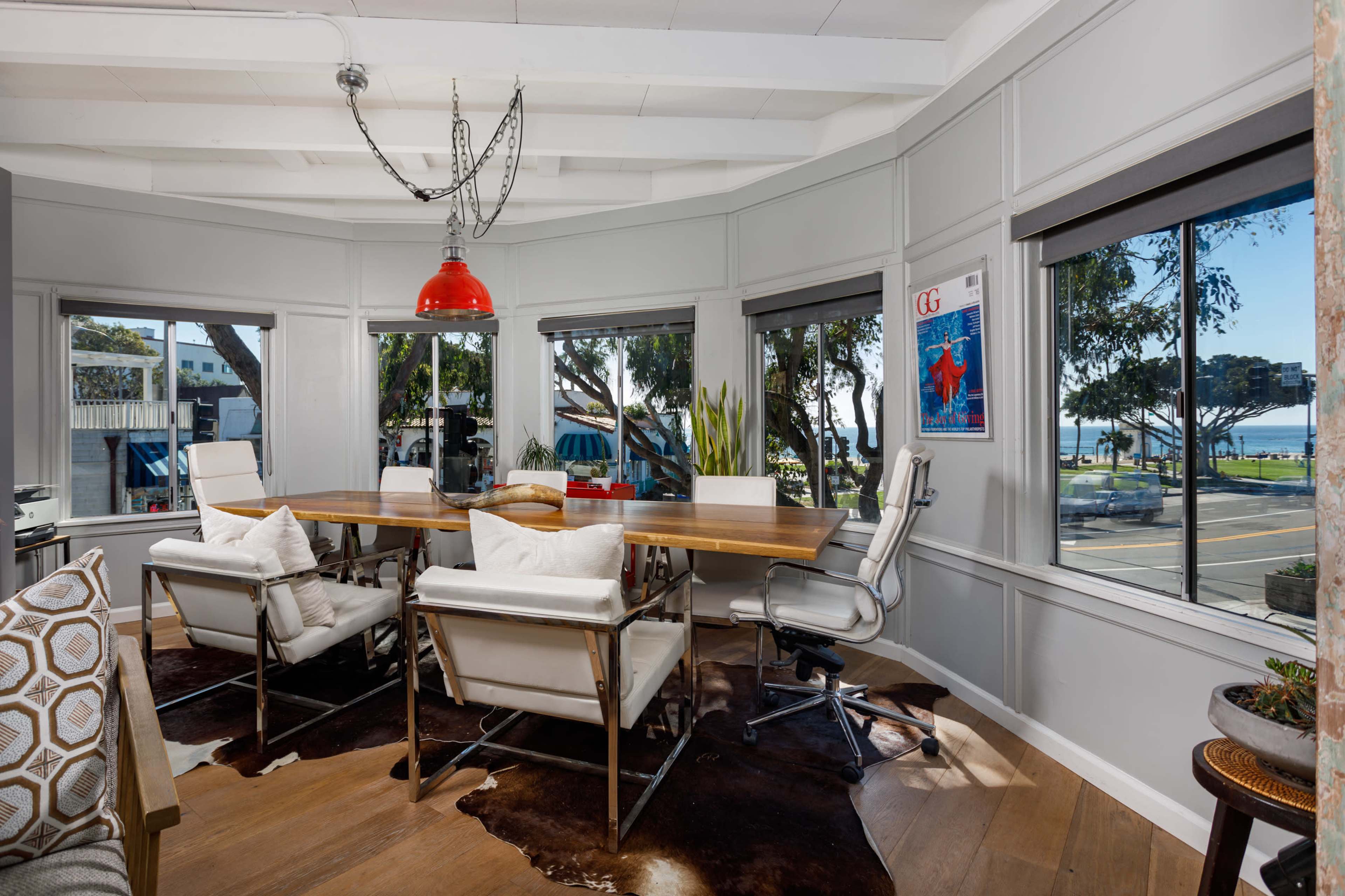 A spacious, sunlit office features a large wooden table surrounded by white chairs and a red pendant light, with windows overlooking a street lined by trees.