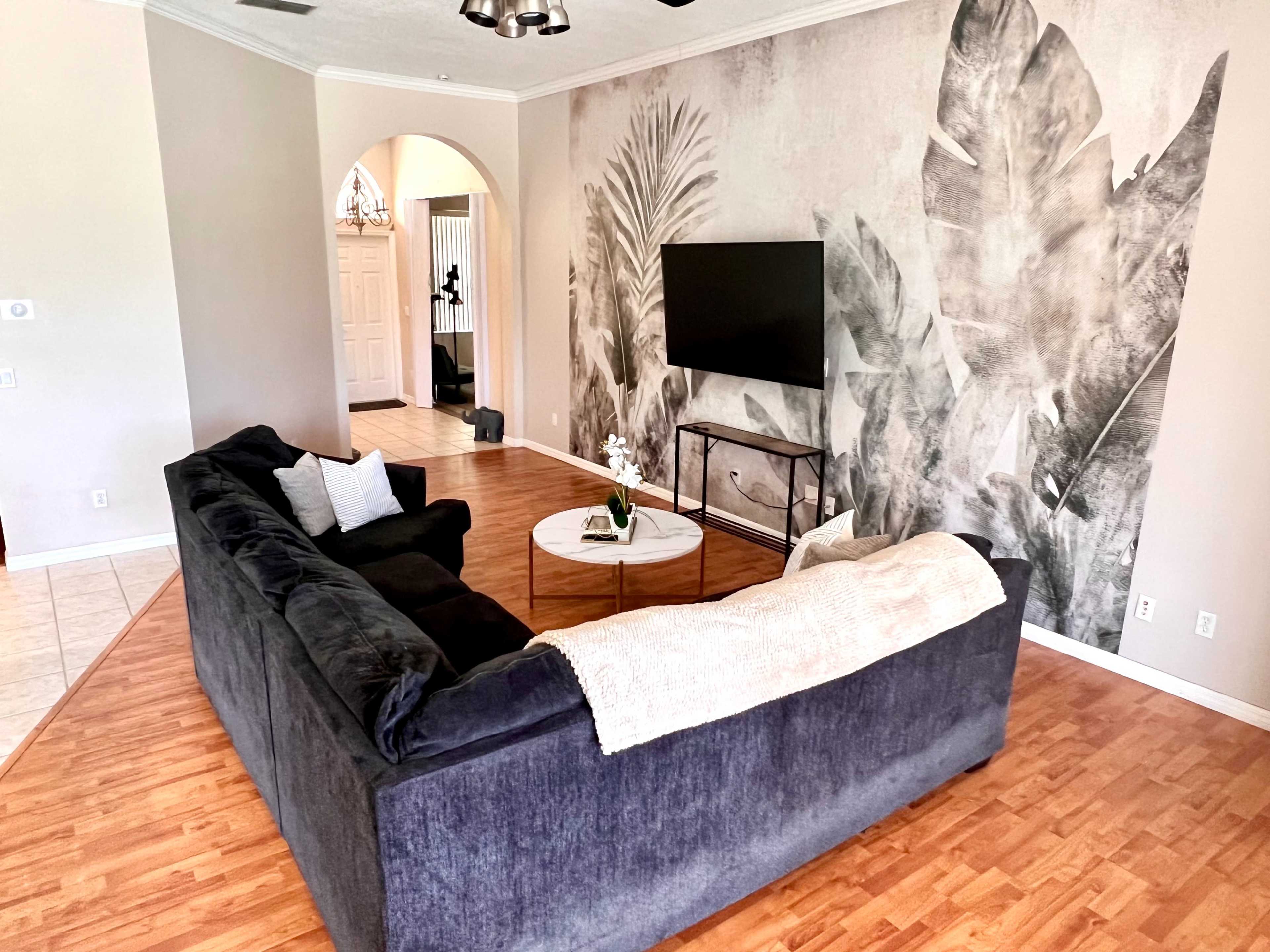 The image depicts a living room featuring a dark sectional sofa, a round coffee table with a plant, and a wall adorned with large, monochromatic leaf prints, along with a television on a metal stand.